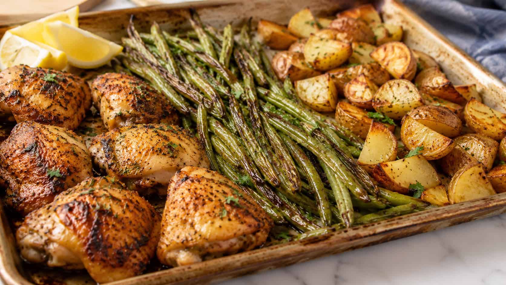 a sheet pan dinner with golden chicken, roasted green beans, and crispy potatoes in a wide horizontal shot, charred edges, sprinkle of herbs, lemon wedges in the corner, bright natural light, shallow depth of field, landscape orientation