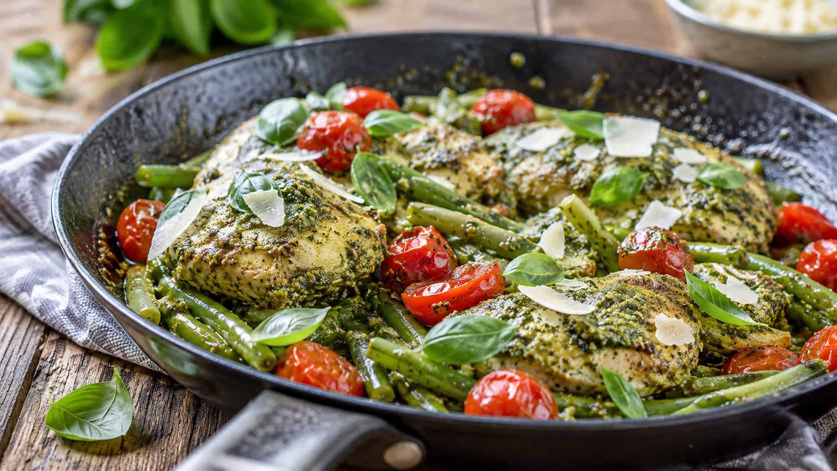 pesto chicken with green beans and burst cherry tomatoes in a skillet in a wide horizontal shot, vibrant green pesto coating, shaved Parmesan and basil on top, rustic table, bright natural light, shallow depth of field, landscape orientation