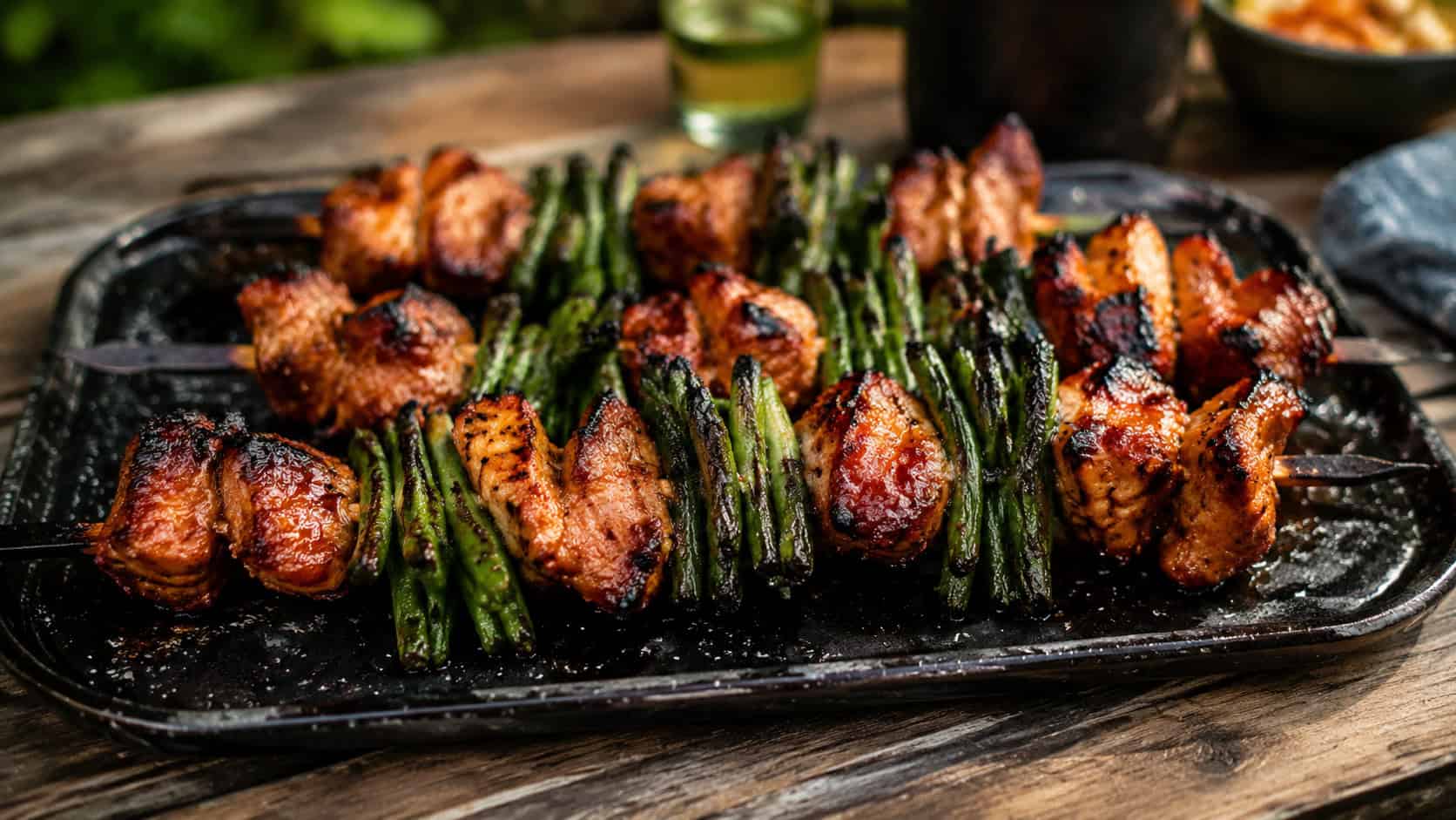 barbecue chicken and green bean kebabs on a grill platter in a wide horizontal shot, sticky charred glaze, blistered green beans, smoky char marks, rustic outdoor table, warm natural light, shallow depth of field, landscape orientation