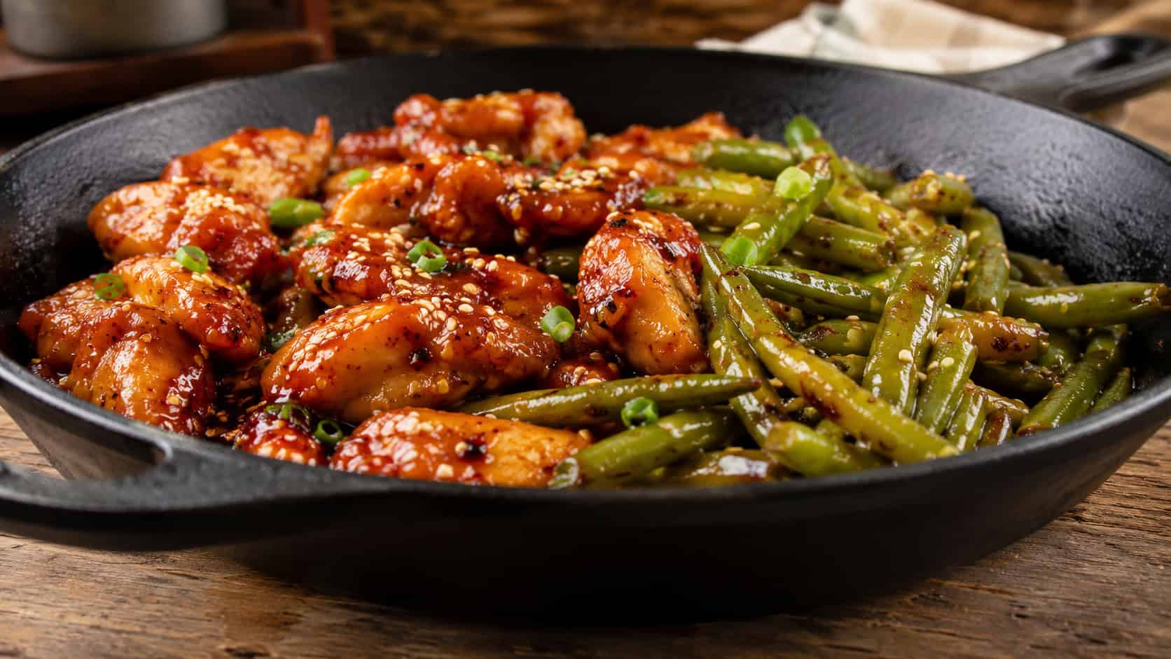 spicy honey chicken and green beans in a cast iron skillet in a wide horizontal shot, glossy sticky glaze with red chili flecks, sesame garnish, rustic wood table, warm natural light, shallow depth of field, landscape orientation