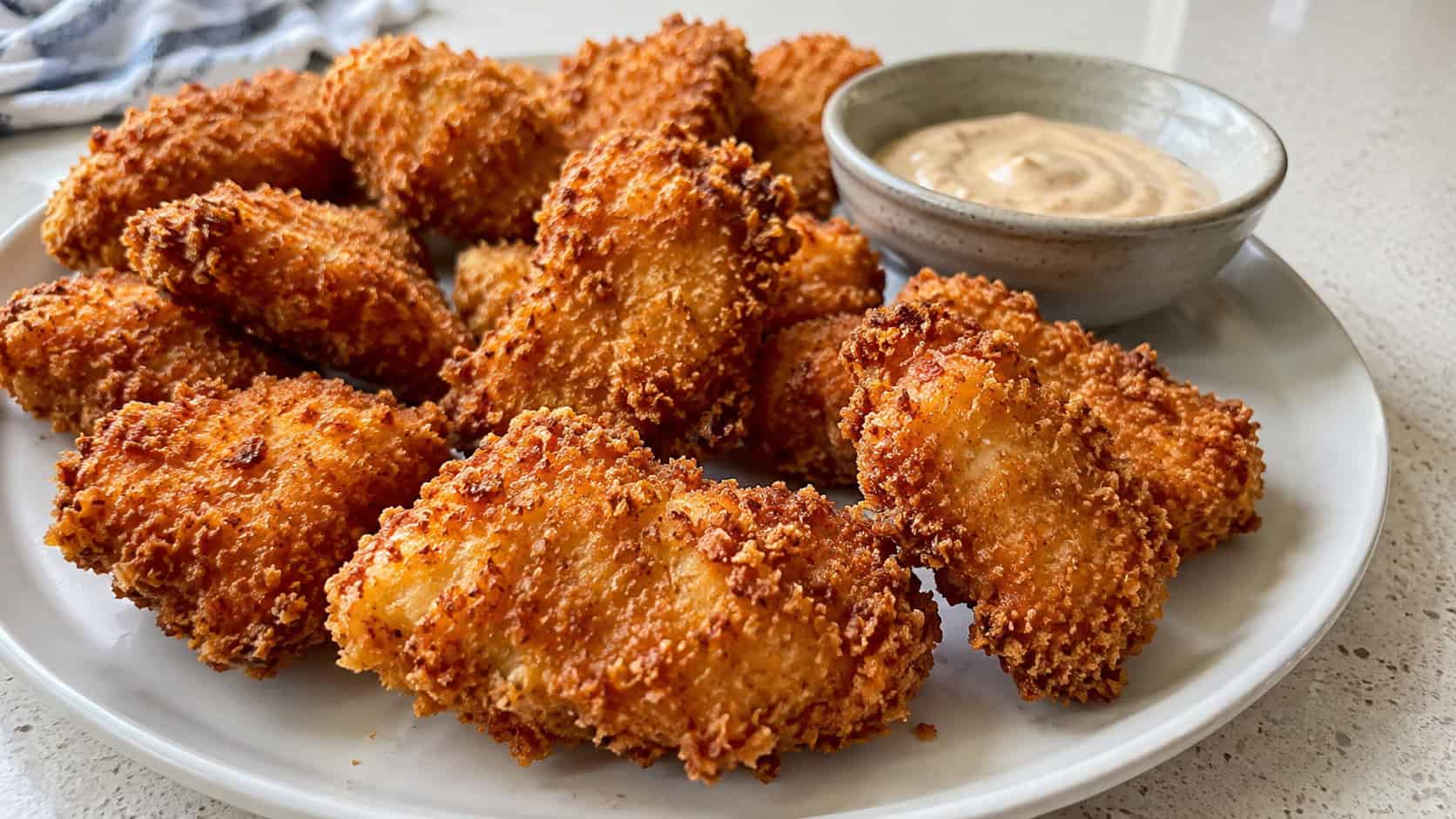 extra-crispy panko chicken nuggets in a wide horizontal shot on a white plate with a craggy golden crust, small dish of dipping sauce, on a bright counter, natural light, shallow depth of field, landscape orientation