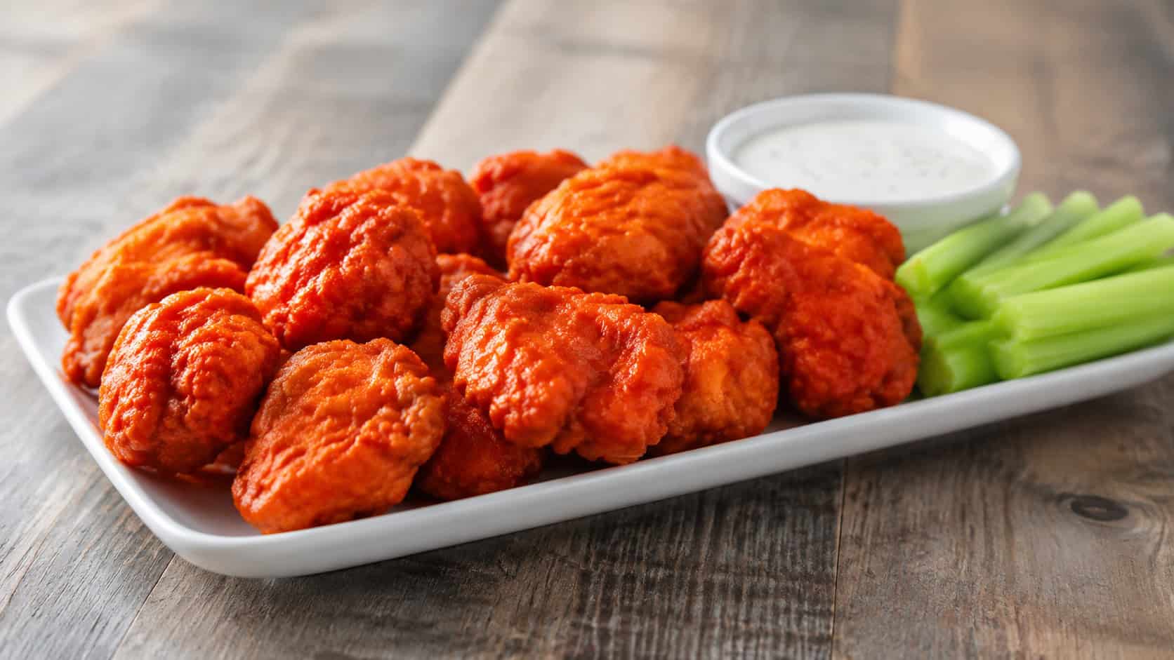 fiery red buffalo chicken nuggets in a wide horizontal shot on a white platter with celery sticks and ranch in a small bowl, rustic wood table, bright natural light, shallow depth of field, landscape orientation