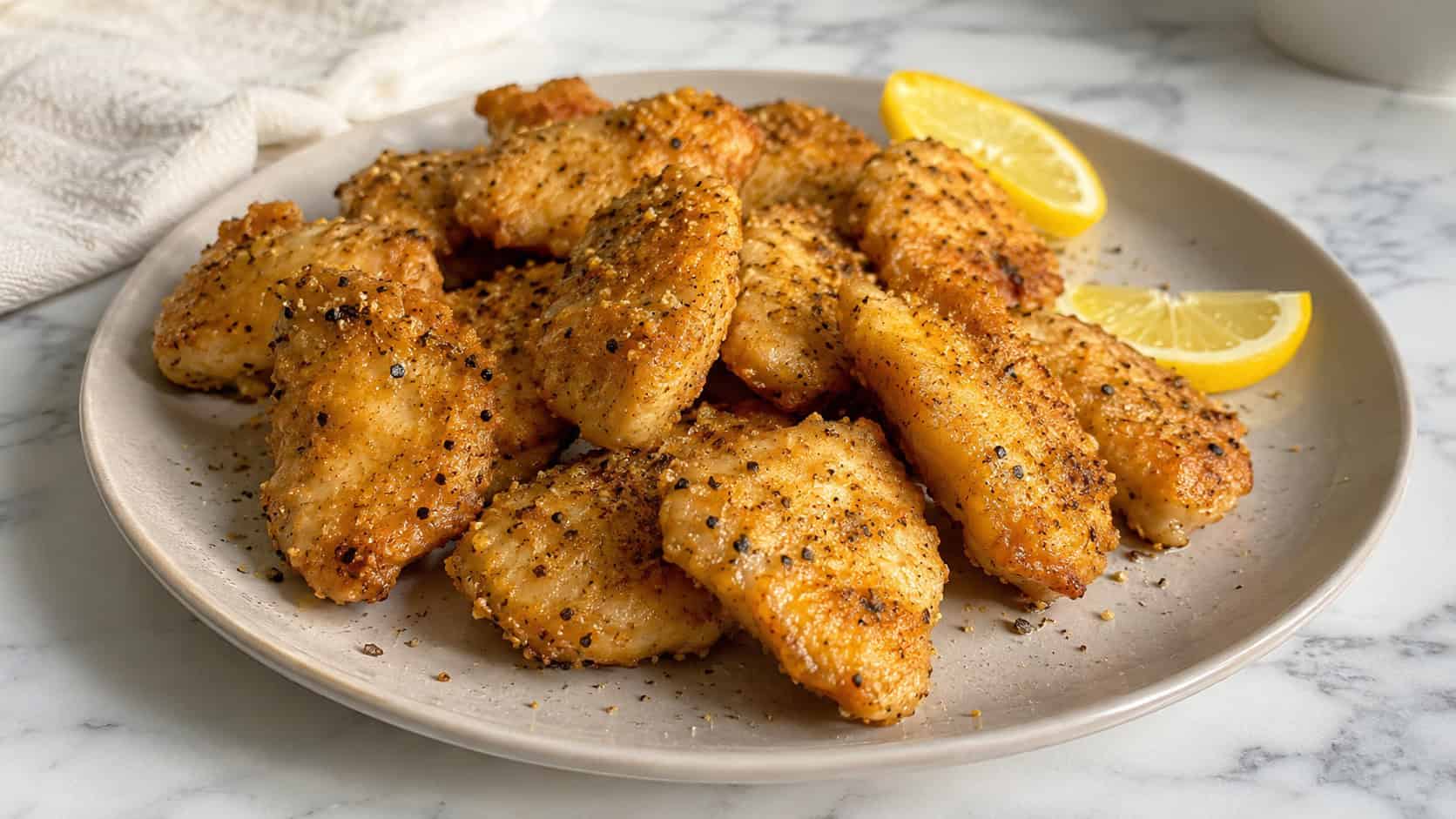 lemon pepper chicken nuggets in a wide horizontal shot on a pale ceramic plate with lemon wedges and cracked pepper, on a bright marble surface, natural light, shallow depth of field, landscape orientation