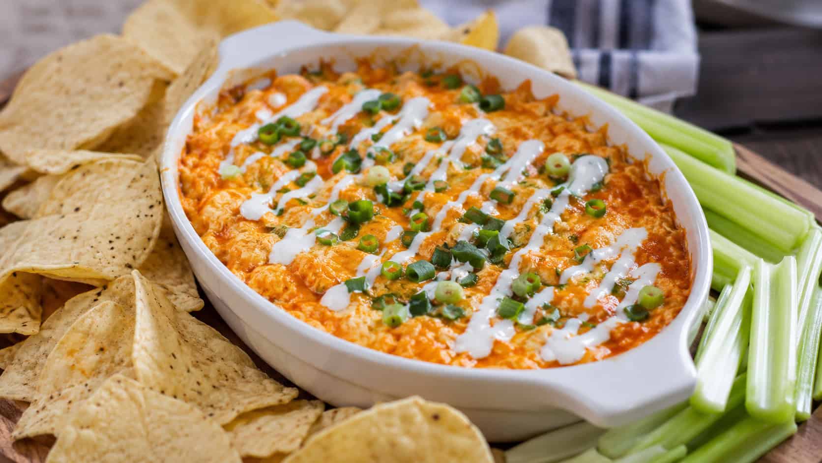 a bubbling golden buffalo chicken dip in a shallow white dish, garnished with chopped green onions and ranch drizzle, surrounded by tortilla chips and celery sticks on a wooden board, wide horizontal shot, natural light, shallow depth of field, landscape orientation