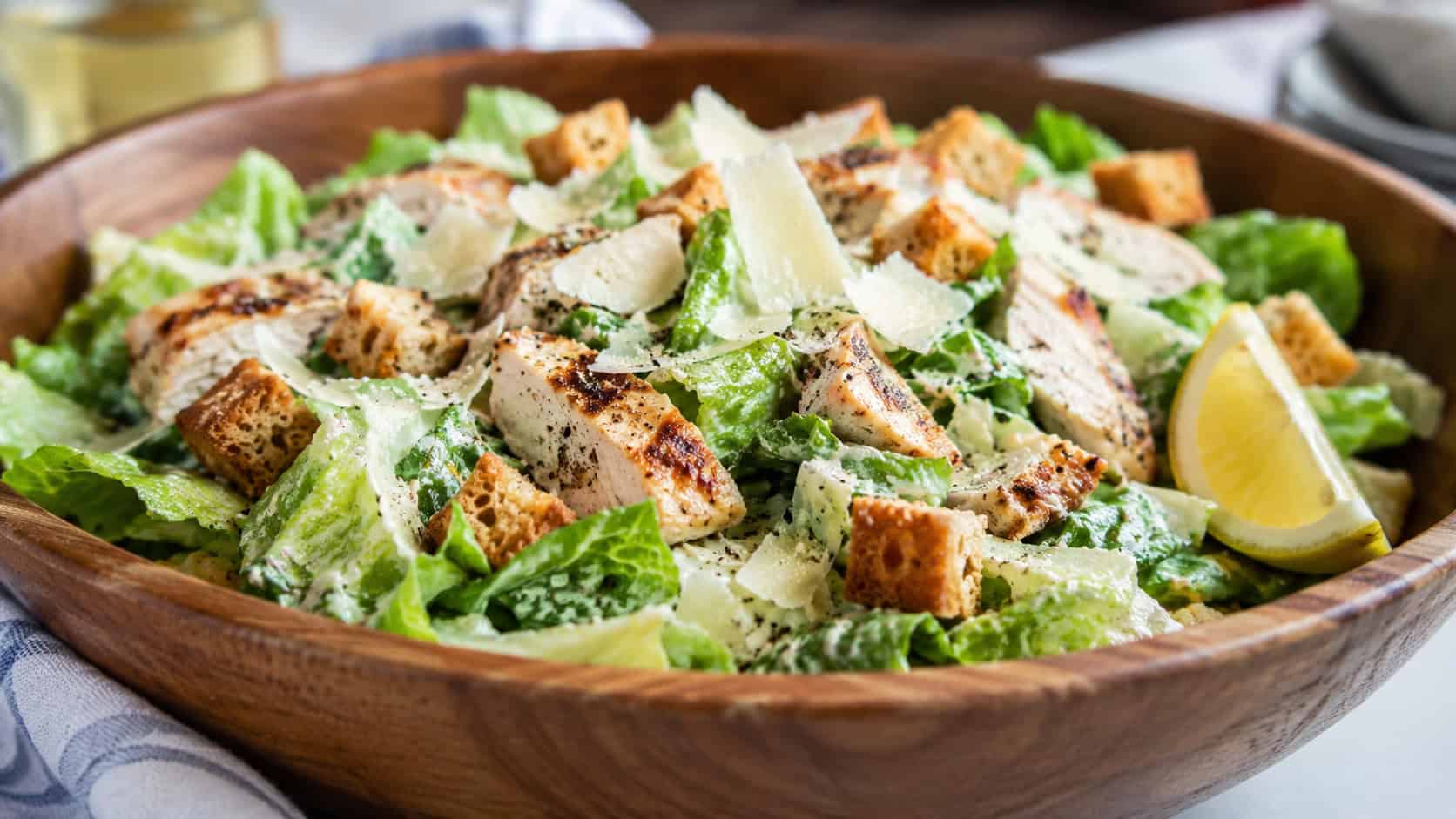 a large chicken Caesar salad with crisp romaine, croutons, and shaved parmesan in a wide wooden bowl, lemon wedge on the side, wide horizontal composition, bright natural light, shallow depth of field, landscape orientation