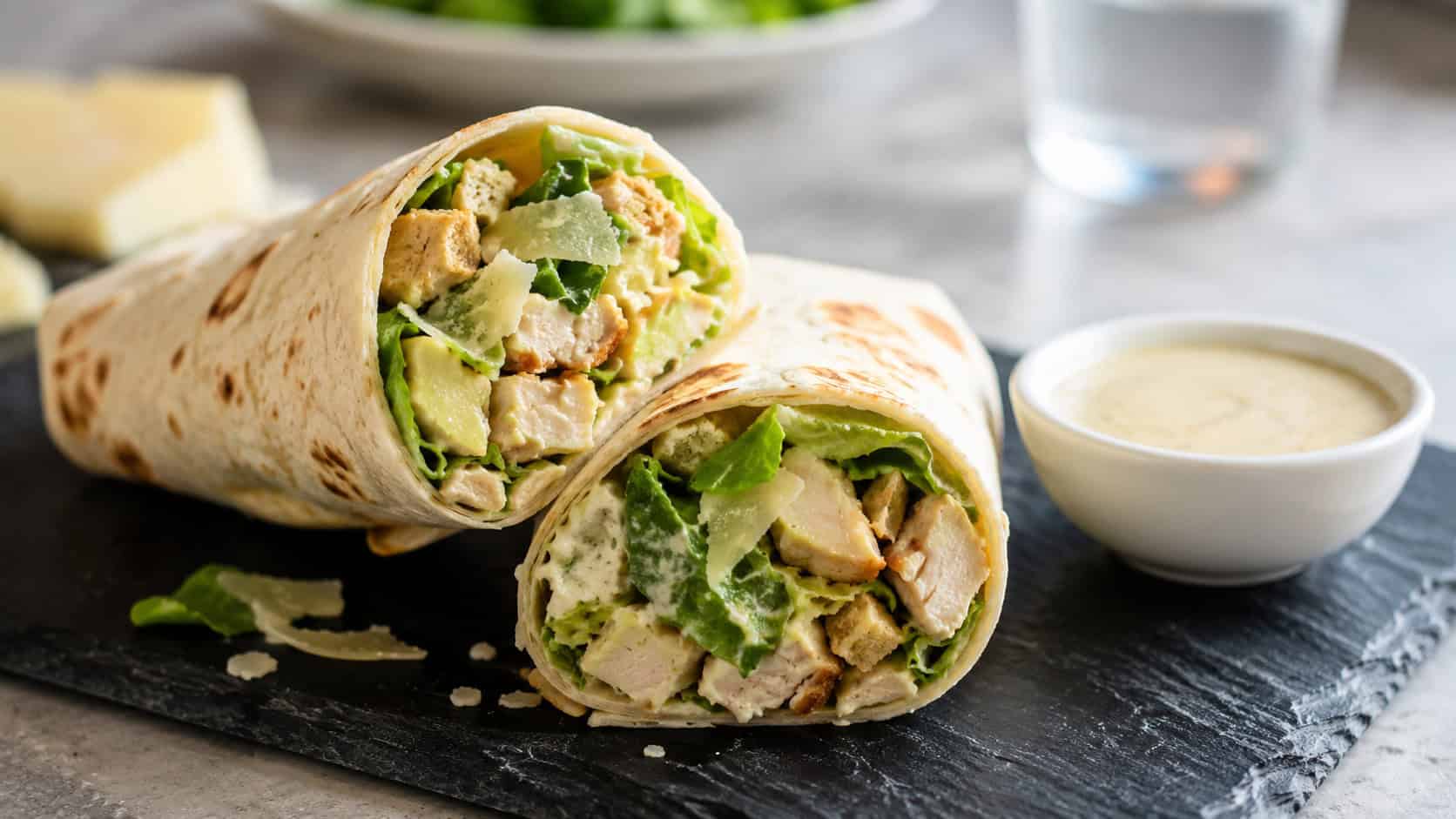 two halved chicken Caesar wraps showing crisp romaine and parmesan inside, arranged on a slate board with a small bowl of dressing, wide horizontal composition, bright natural light, shallow depth of field, landscape orientation