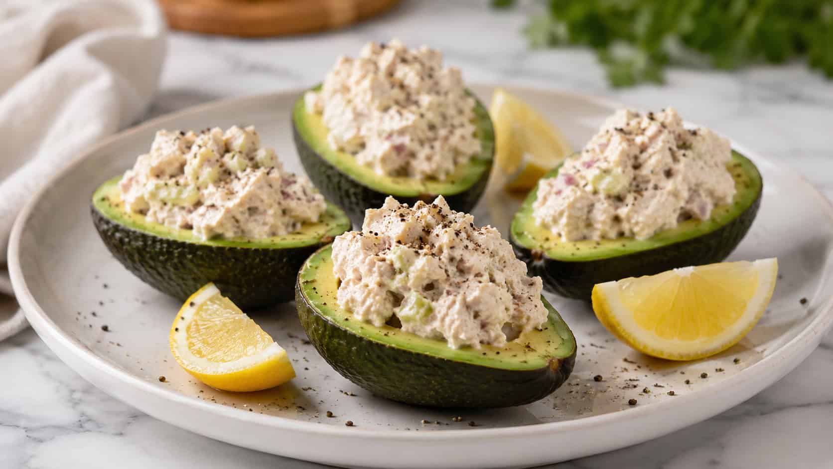 four avocado halves filled with creamy chicken salad and topped with cracked pepper, plated on a marble surface with lemon wedges scattered around, wide horizontal shot, soft natural light, shallow depth of field, landscape orientation