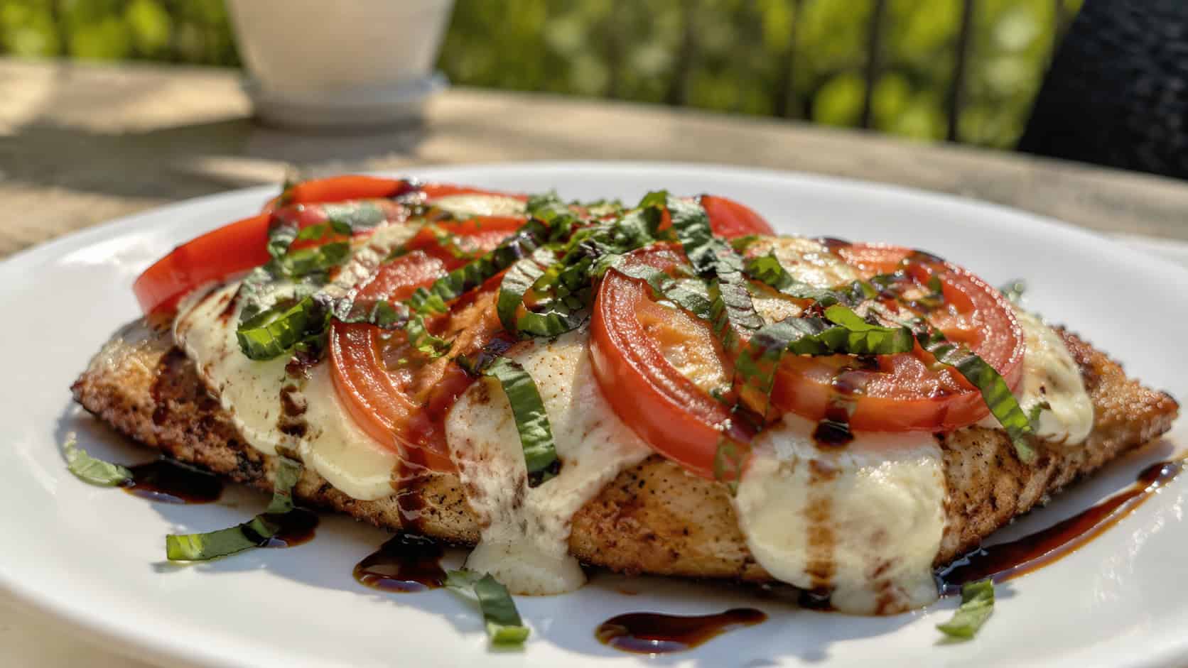 chicken caprese topped with melted mozzarella, tomato and basil drizzled with balsamic, on a white plate on a sunny table, natural light, shallow depth of field, wide horizontal landscape composition