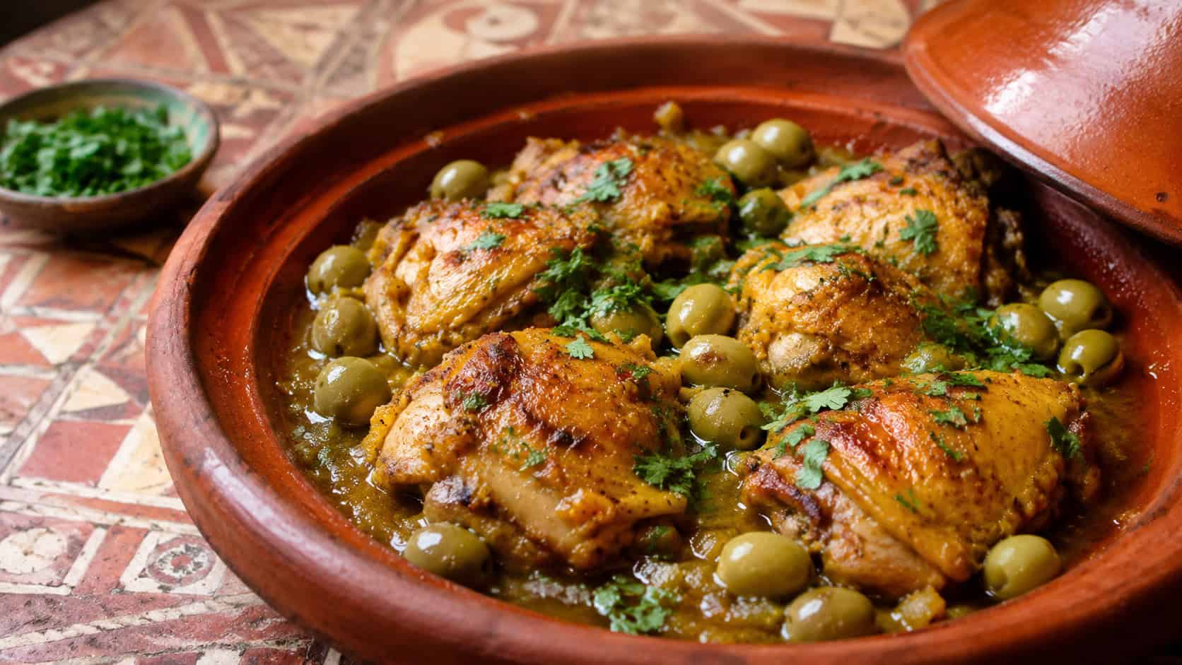 golden braised chicken thighs with green olives in a rustic clay tagine, garnished with cilantro, on a warm patterned table, natural light, shallow depth of field, wide horizontal landscape composition