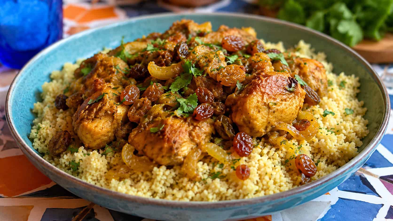 spiced Moroccan chicken with raisins over a mound of fluffy couscous in a wide bowl, sprinkled with herbs, on a colorful tabletop, natural light, shallow depth of field, wide horizontal landscape composition