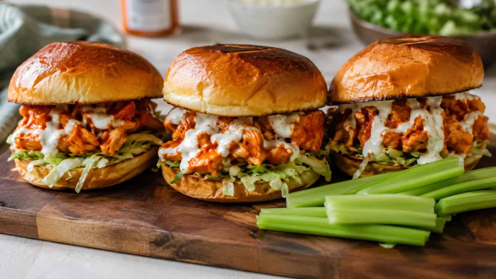 a wide horizontal plate of buffalo chicken sandwiches on toasted buns with lettuce and ranch drizzle, celery on the side, on a wood board, natural light, shallow depth of field, landscape orientation