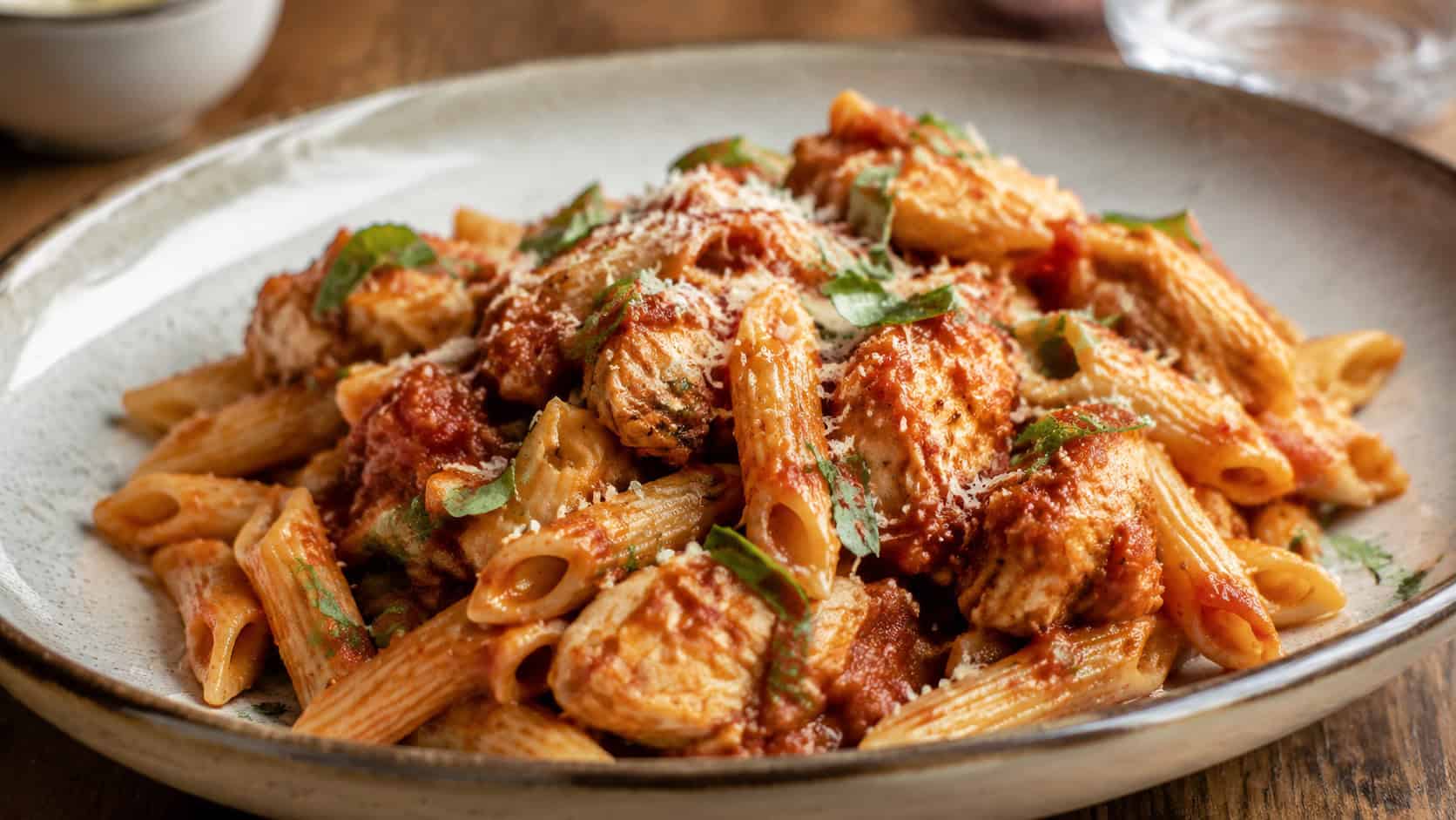 a wide horizontal bowl of saucy Italian chicken penne with Parmesan and basil, rich red sauce, on a ceramic plate, warm natural light, shallow depth of field, landscape orientation