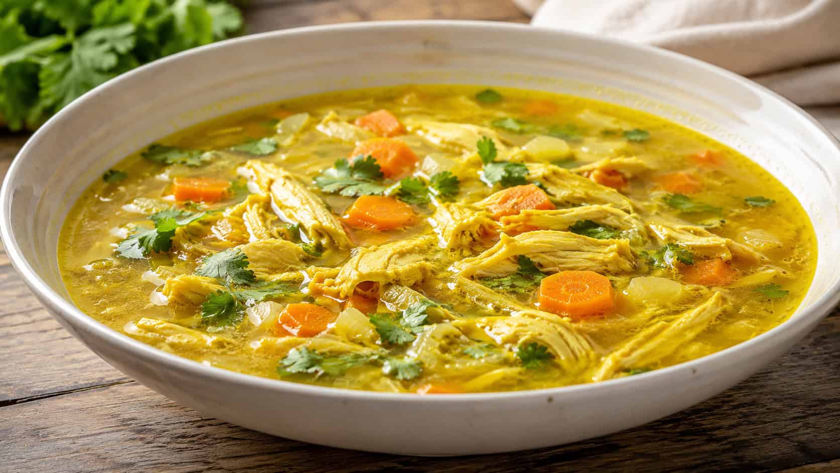 a wide horizontal bowl of golden turmeric chicken soup with carrots and shredded chicken, cilantro garnish, warm glowing broth, natural light, shallow depth of field, landscape orientation