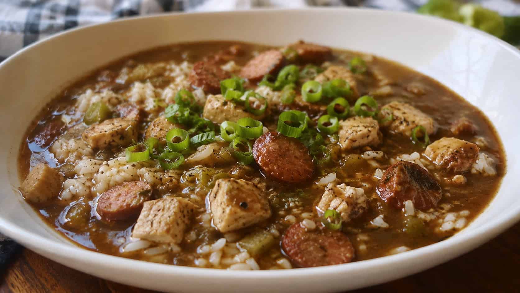 a wide horizontal bowl of Cajun chicken and sausage gumbo over white rice, deep brown broth with sausage, scallions on top, natural light, shallow depth of field, landscape orientation