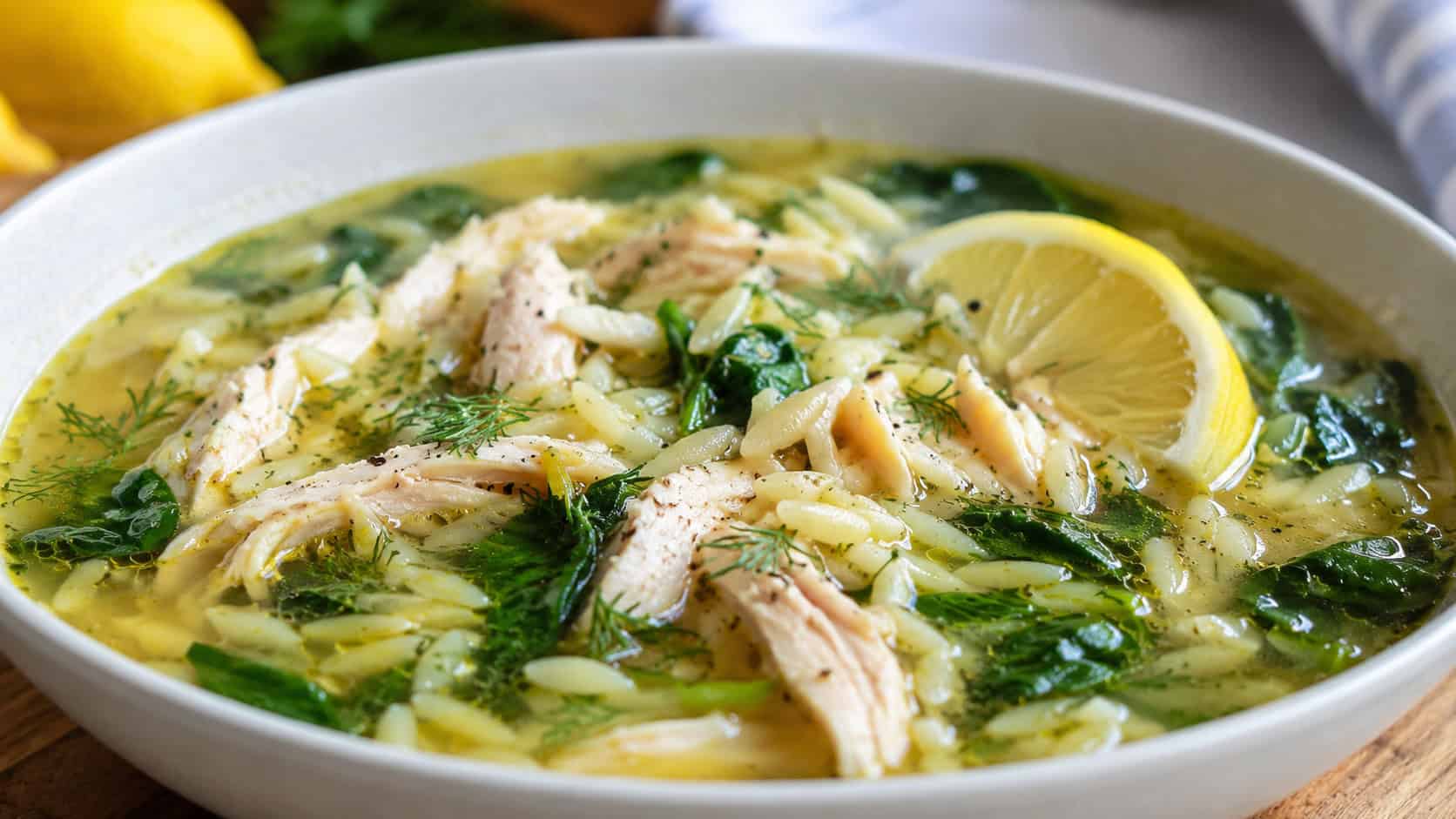 a wide horizontal bowl of lemon chicken orzo soup with spinach and a lemon wedge, bright broth and fresh dill, natural light, shallow depth of field, landscape orientation