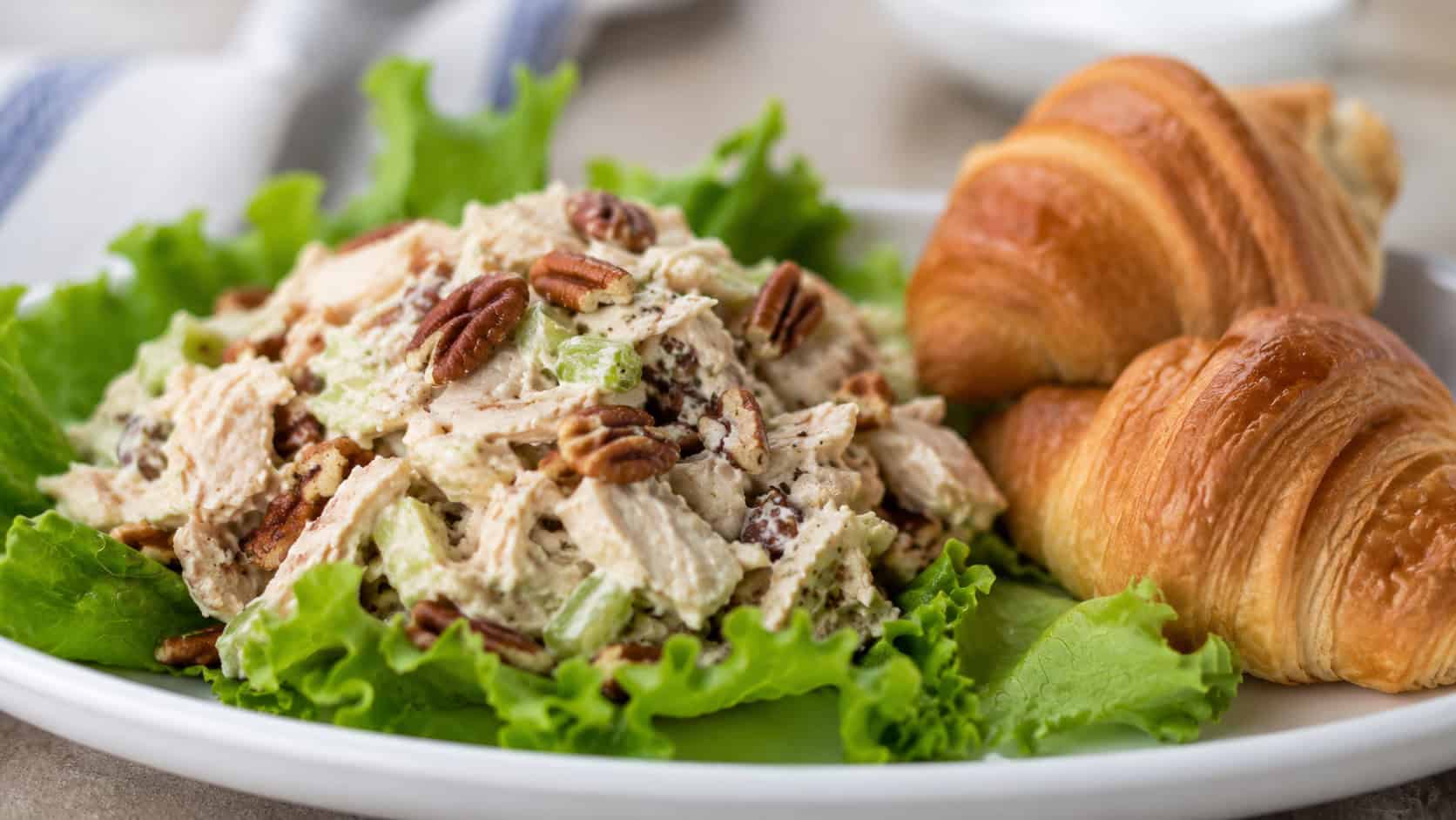 creamy chicken pecan salad mounded on a leaf of lettuce in a wide horizontal shot, toasted pecans visible, croissants beside it, bright clean plate, soft natural light, shallow depth of field, landscape orientation