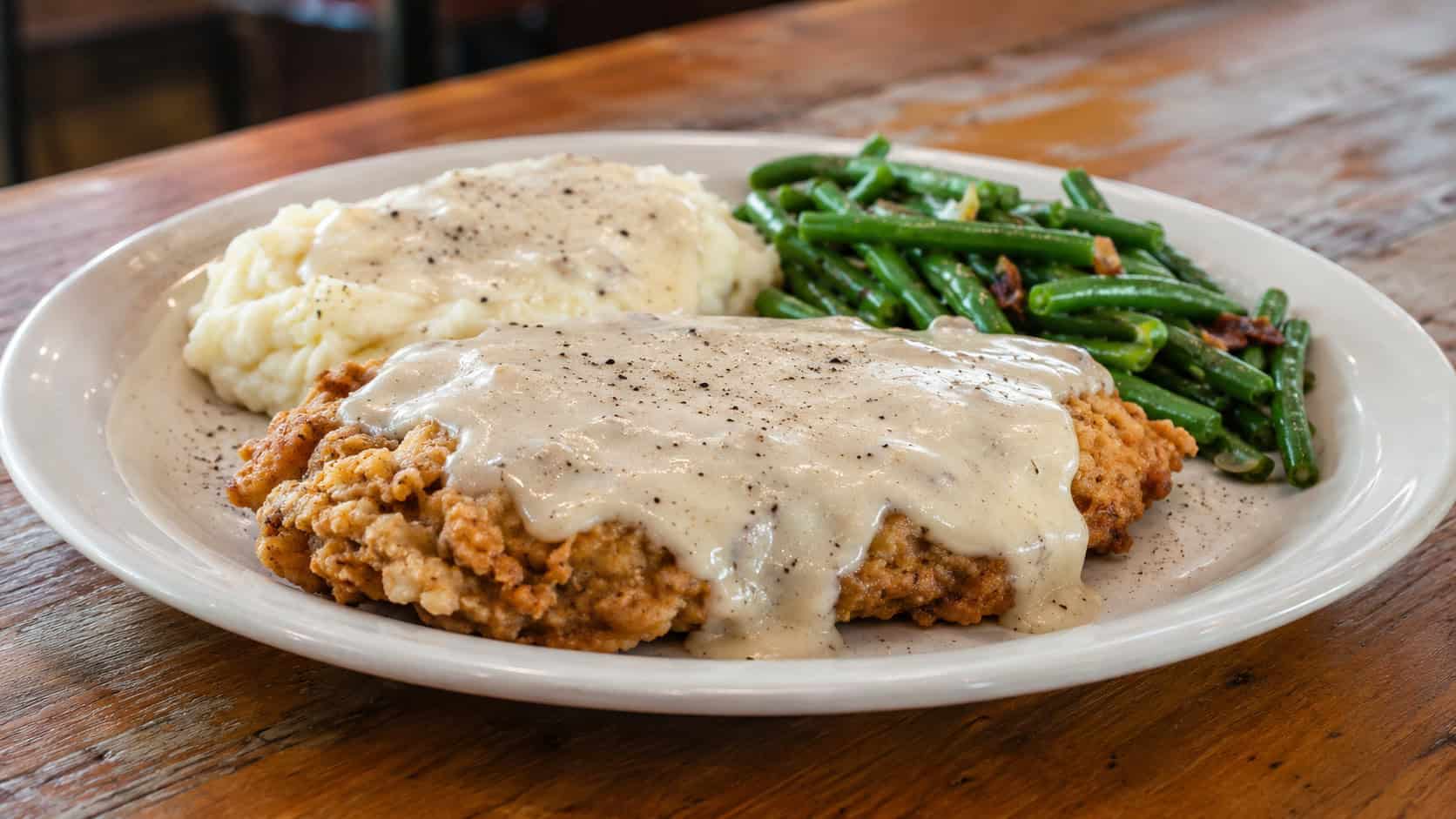 crispy chicken-fried chicken breast smothered in creamy peppered gravy on a plate in a wide horizontal shot, mashed potatoes and green beans beside it, rustic diner table, warm natural light, shallow depth of field, landscape orientation