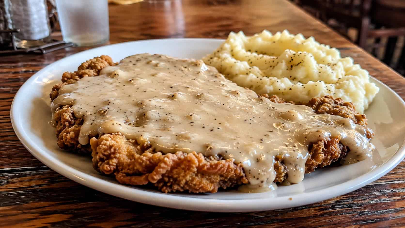 crispy chicken fried steak smothered in creamy peppered gravy on a plate in a wide horizontal shot, mashed potatoes beside it, rustic diner table, warm natural light, shallow depth of field, landscape orientation