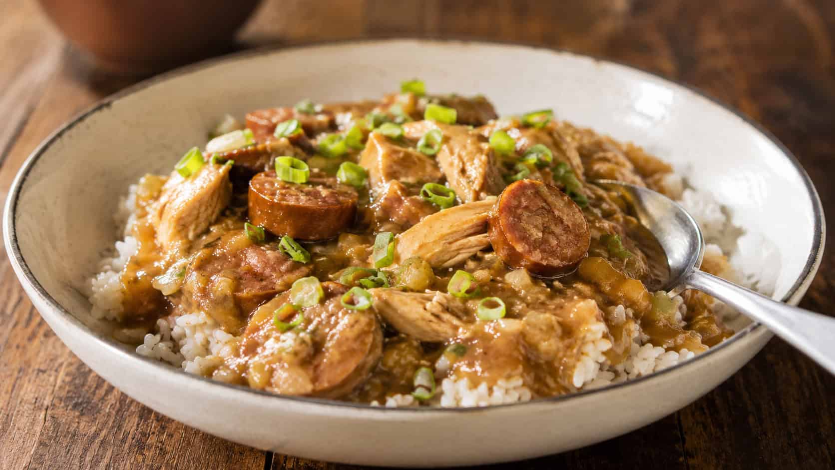 chicken and sausage étouffée in a thick golden sauce over white rice in a wide horizontal shot, green onions on top, a spoon resting in the bowl, rustic table, warm natural light, shallow depth of field, landscape orientation