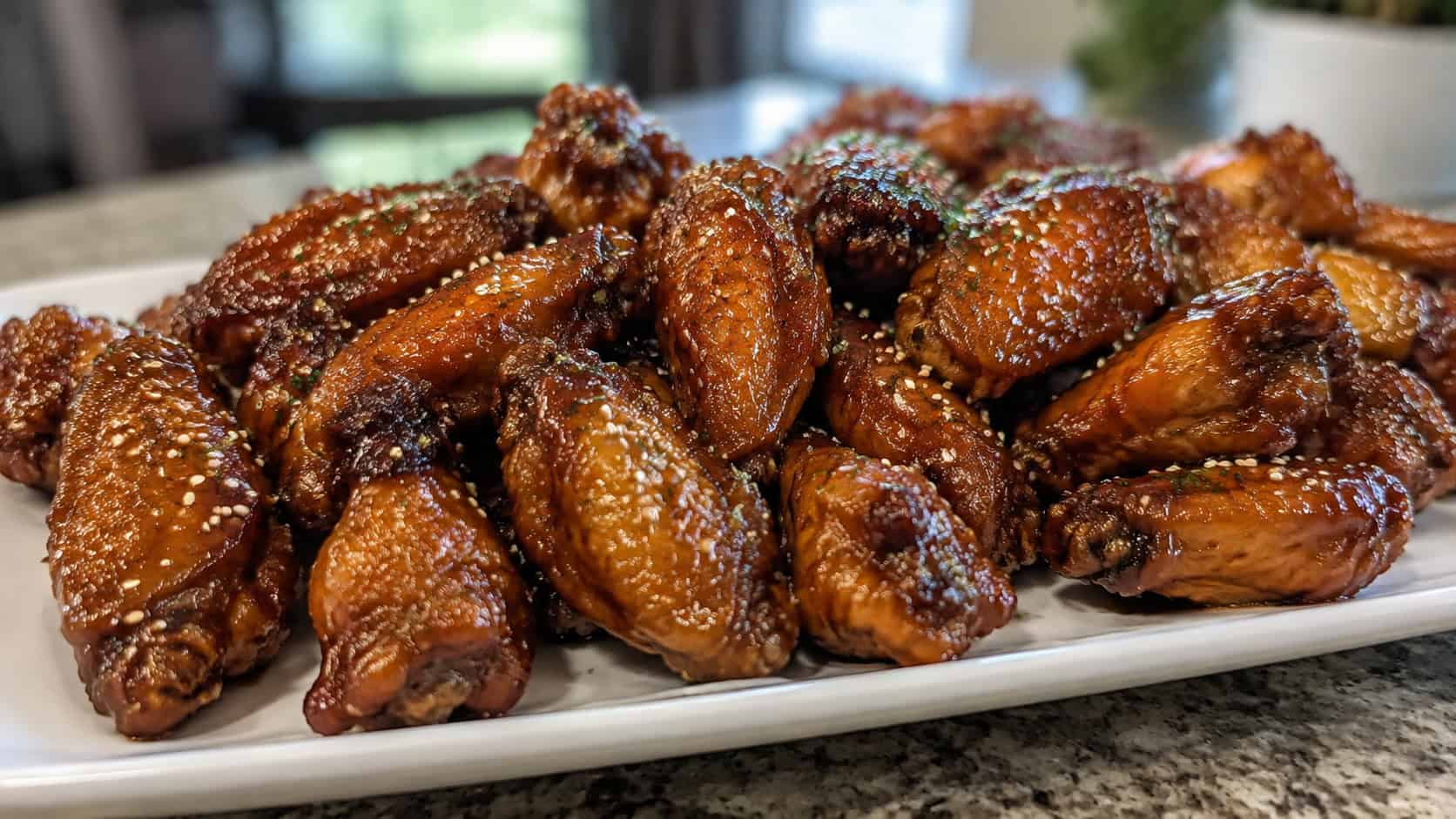 glossy honey garlic chicken wings sprinkled with sesame seeds piled on a wide platter, set on a counter, natural light, shallow depth of field, landscape orientation