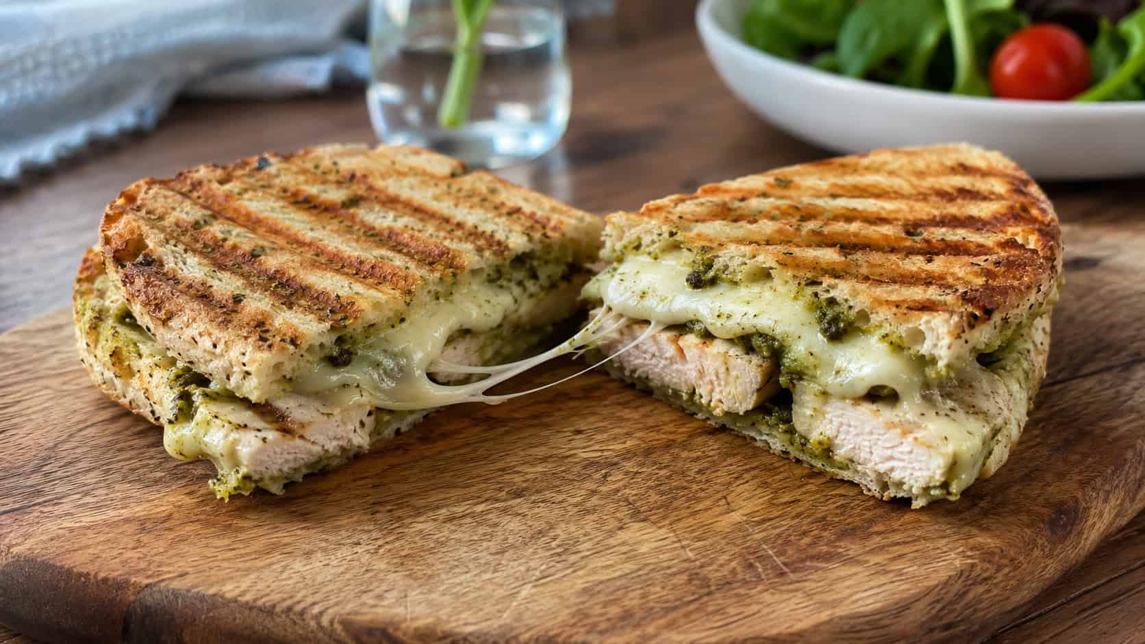 a pressed pesto chicken panini cut in half showing melted mozzarella and grilled chicken on a wide board, set on a table, natural light, shallow depth of field, landscape orientation