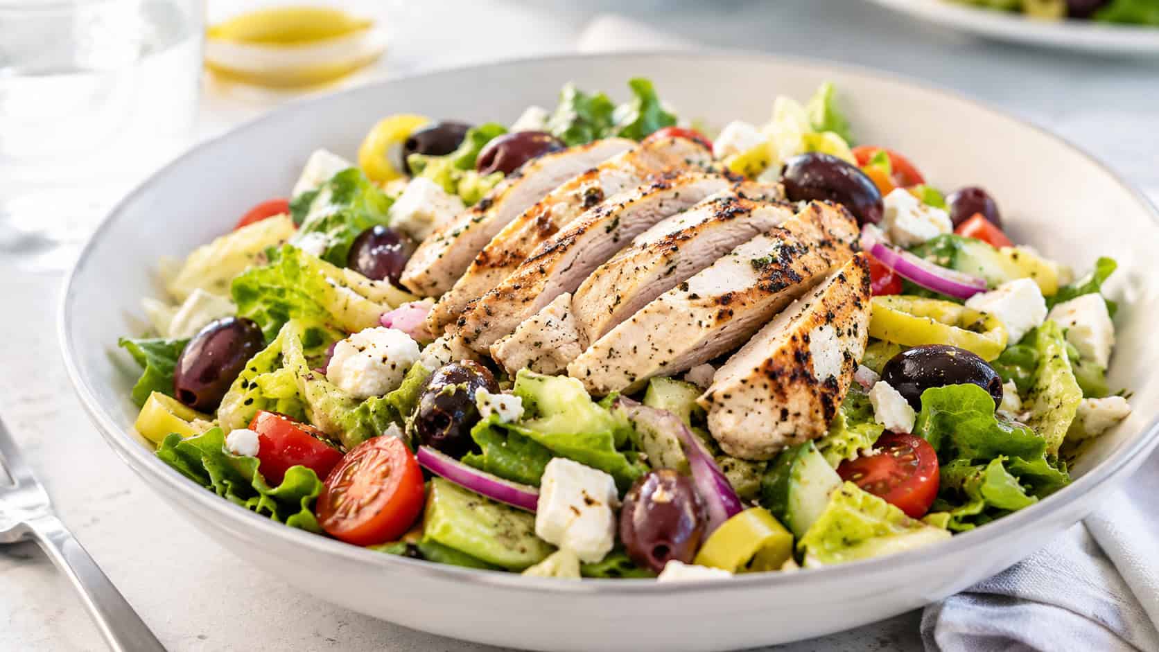 a Greek chicken salad with grilled chicken, feta, and olives over crisp greens in a wide shallow bowl, set on a bright table, natural light, shallow depth of field, landscape orientation