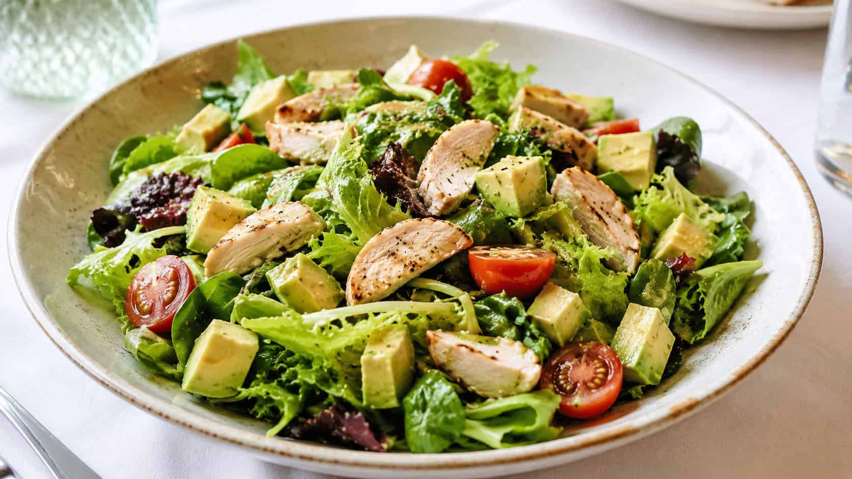 a fresh chicken and avocado salad with green leaves, cubed avocado, and cherry tomatoes in a wide horizontal shot, served in a wide shallow bowl on a white tablecloth, crisp bright natural light, shallow depth of field, landscape orientation