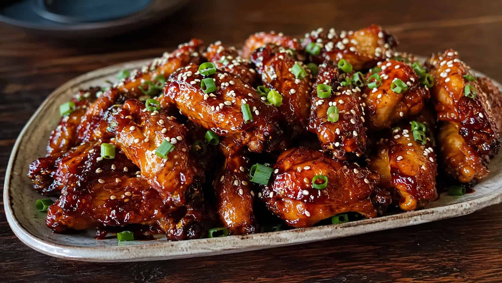 sticky ginger soy chicken wings glistening with glaze in a wide horizontal shot, piled on a platter and sprinkled with sesame seeds and green onion, set on a dark wood table, warm natural light, shallow depth of field, landscape orientation
