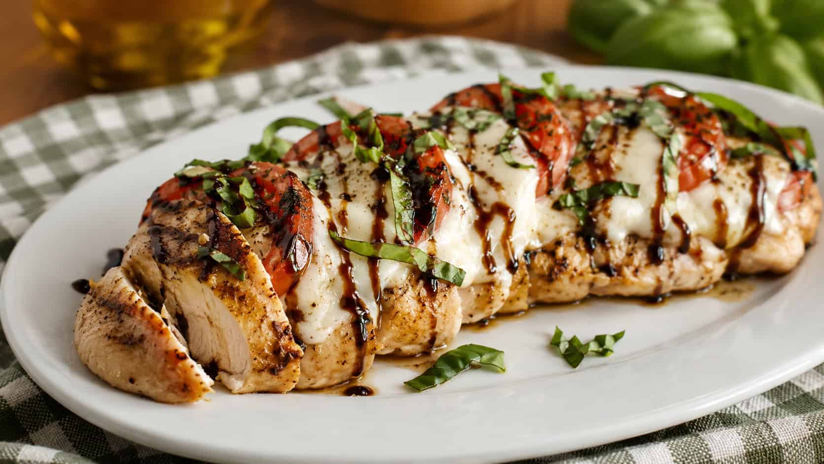 Italian chicken caprese topped with melted mozzarella, tomato slices, and fresh basil in a wide horizontal shot, drizzled with dark balsamic on a white plate over a checkered cloth, warm natural light, shallow depth of field, landscape orientation