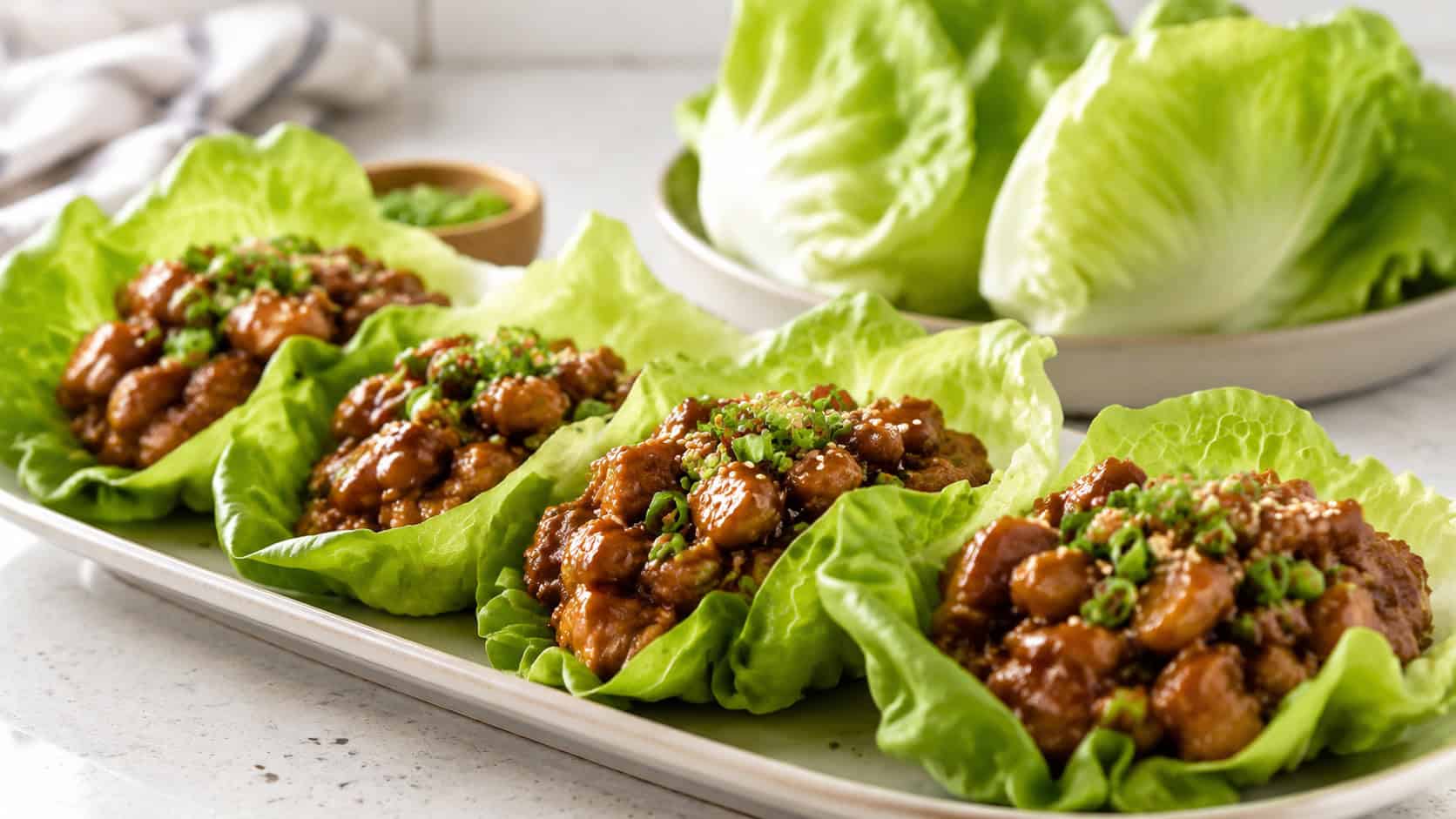 teriyaki chicken lettuce wraps filled with saucy chicken and topped with green onion in a wide horizontal shot, arranged on a platter with extra lettuce cups on a bright counter, fresh natural light, shallow depth of field, landscape orientation