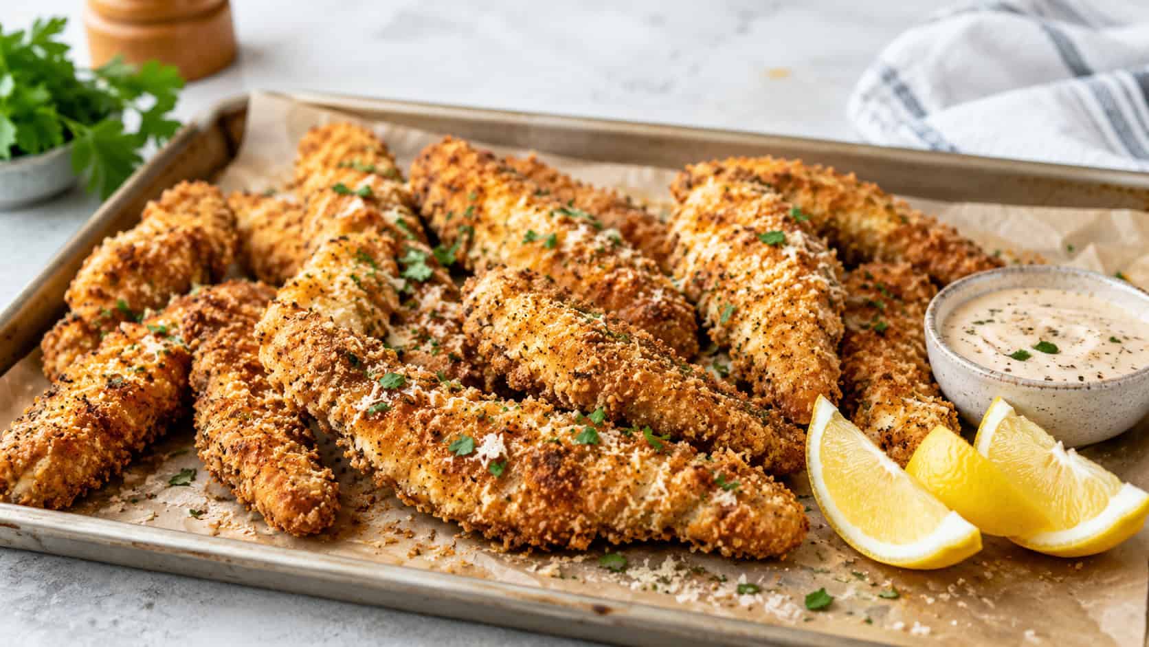 golden crispy garlic parmesan chicken tenders piled on a parchment-lined sheet pan in a wide horizontal shot, small bowl of dip and lemon wedges beside them on a light kitchen counter, bright natural light, shallow depth of field, landscape orientation