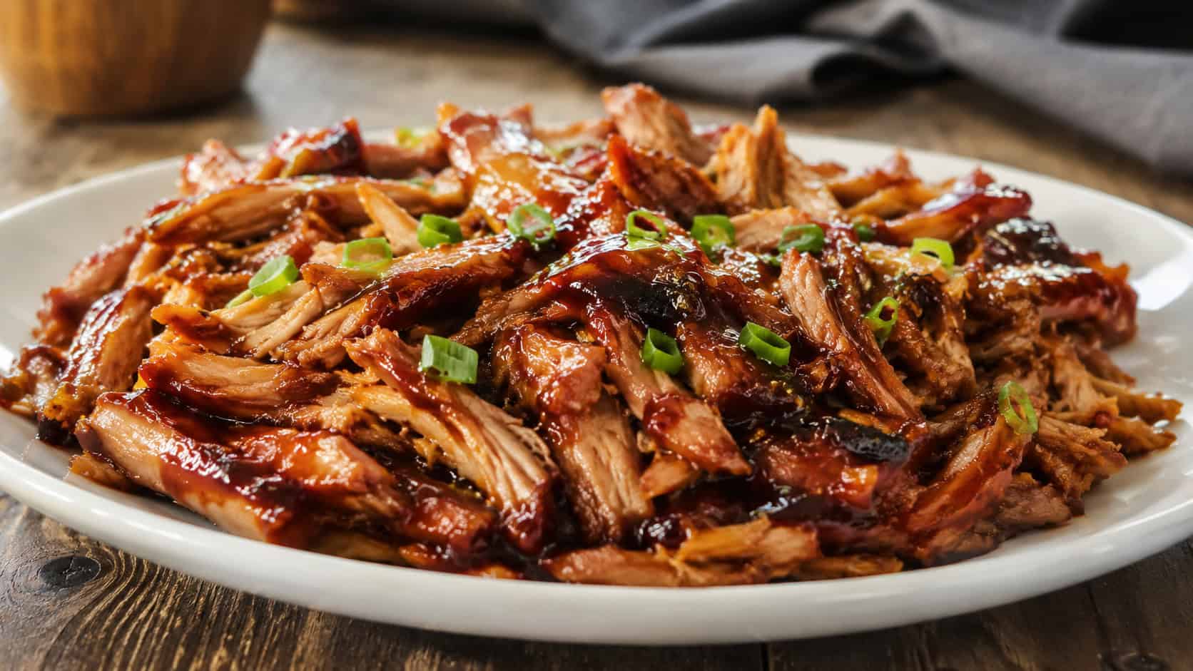 saucy BBQ honey garlic chicken shredded and piled in a wide horizontal frame on a white plate, glossy caramelized sauce with a sprinkle of green onion, rustic wood table, warm natural light, shallow depth of field, landscape orientation