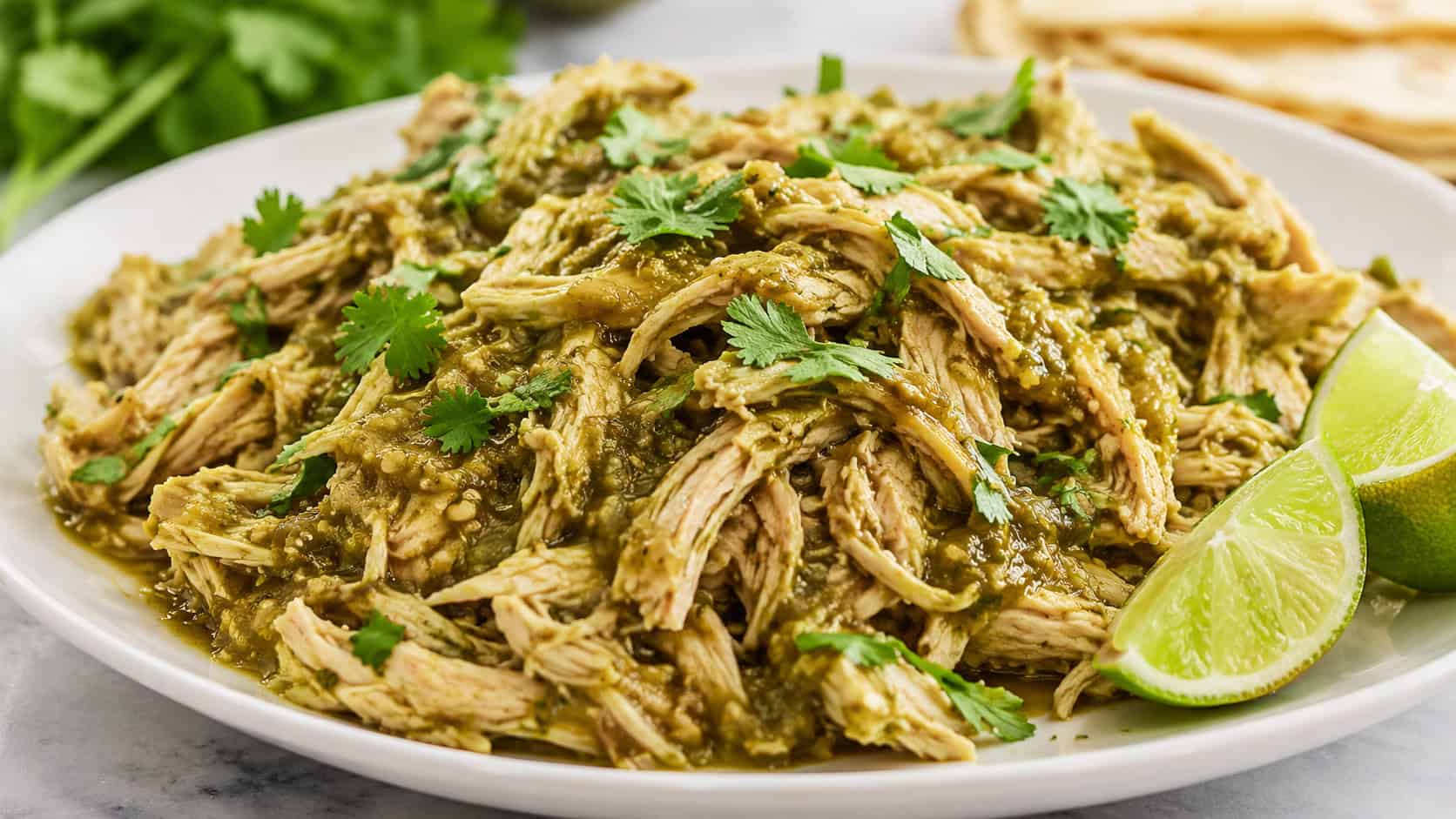 salsa verde shredded chicken in a wide horizontal frame on a plate, tender chicken coated in green salsa topped with cilantro and lime wedges, bright fresh light, shallow depth of field, landscape orientation