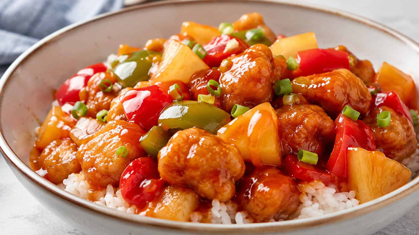 sweet and sour chicken in a wide horizontal frame in a bowl, glossy red-orange sauce with chunks of chicken bell pepper and pineapple over rice, bright clean light, shallow depth of field, landscape orientation