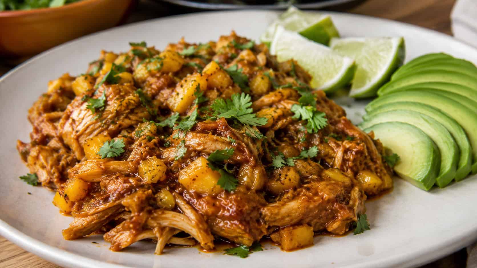 mango chipotle shredded chicken in a wide horizontal frame on a plate, saucy chicken with flecks of mango topped with cilantro and lime, avocado slices nearby, bright warm light, shallow depth of field, landscape orientation