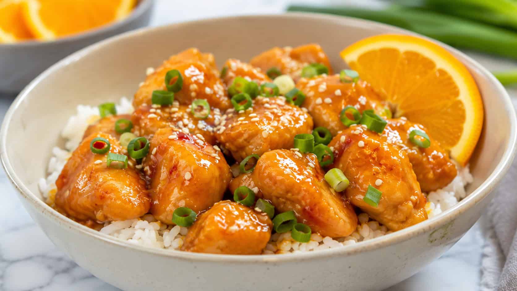 tangy orange chicken over rice in a wide horizontal frame in a bowl, glossy orange-glazed chicken pieces with sesame seeds and green onion, orange slice garnish, bright clean light, shallow depth of field, landscape orientation