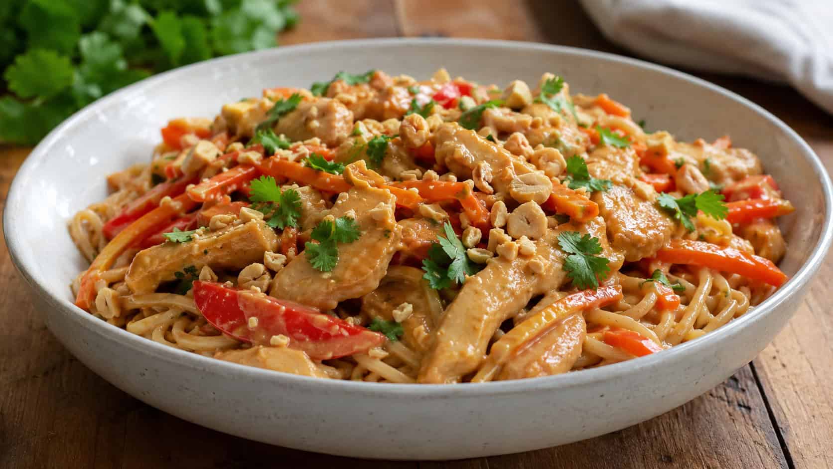 peanut sauce chicken stir fry with bell pepper and shredded carrots in a wide horizontal shot, creamy tan sauce coating everything, served over noodles in a wide bowl, topped with crushed peanuts and cilantro, on a wooden table, natural light, shallow depth of field, landscape orientation