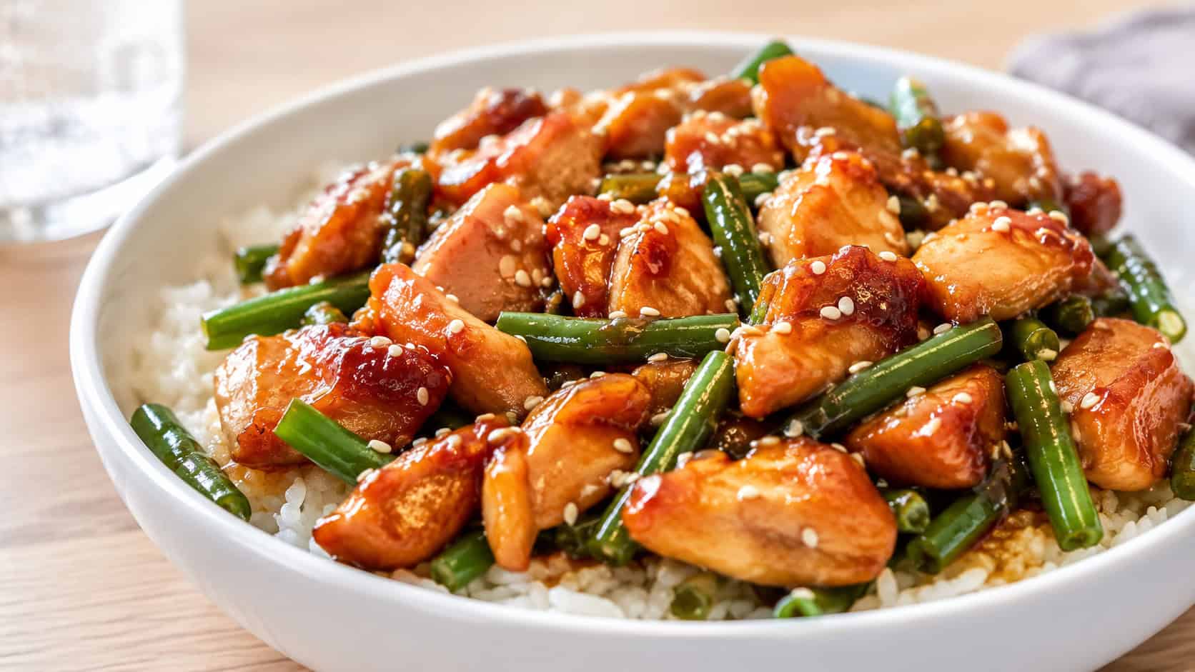honey garlic chicken stir fry with green beans in a wide horizontal shot, sticky golden glaze coating the chicken, served in a white bowl over rice, sprinkled with sesame seeds, on a bright wooden table, natural light, shallow depth of field, landscape orientation