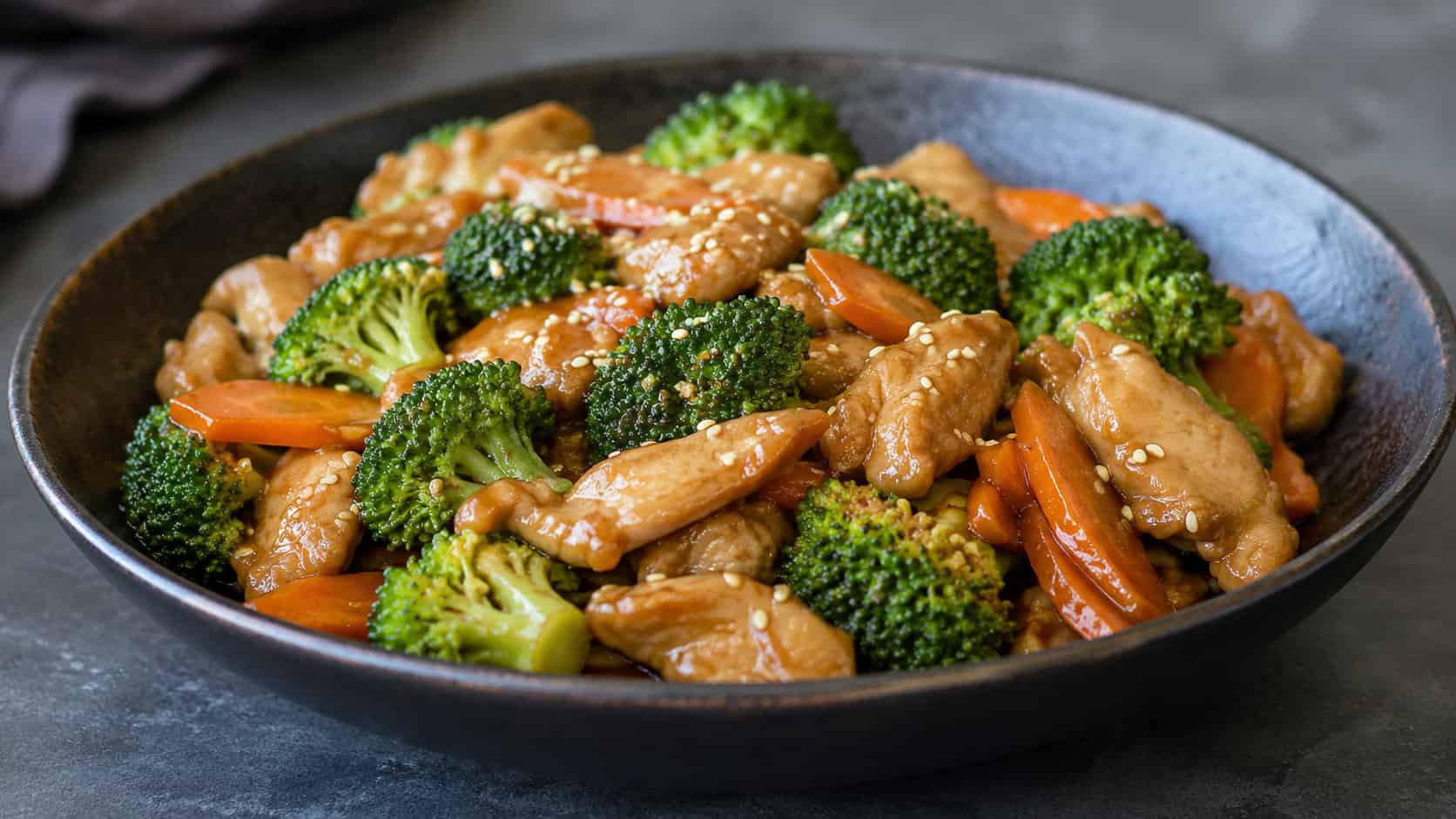 garlic ginger chicken stir fry with broccoli and sliced carrots in a wide horizontal shot, piled in a dark ceramic bowl, glossy sauce coating the chicken, sesame seeds scattered on top, on a slate gray surface, natural light, shallow depth of field, landscape orientation
