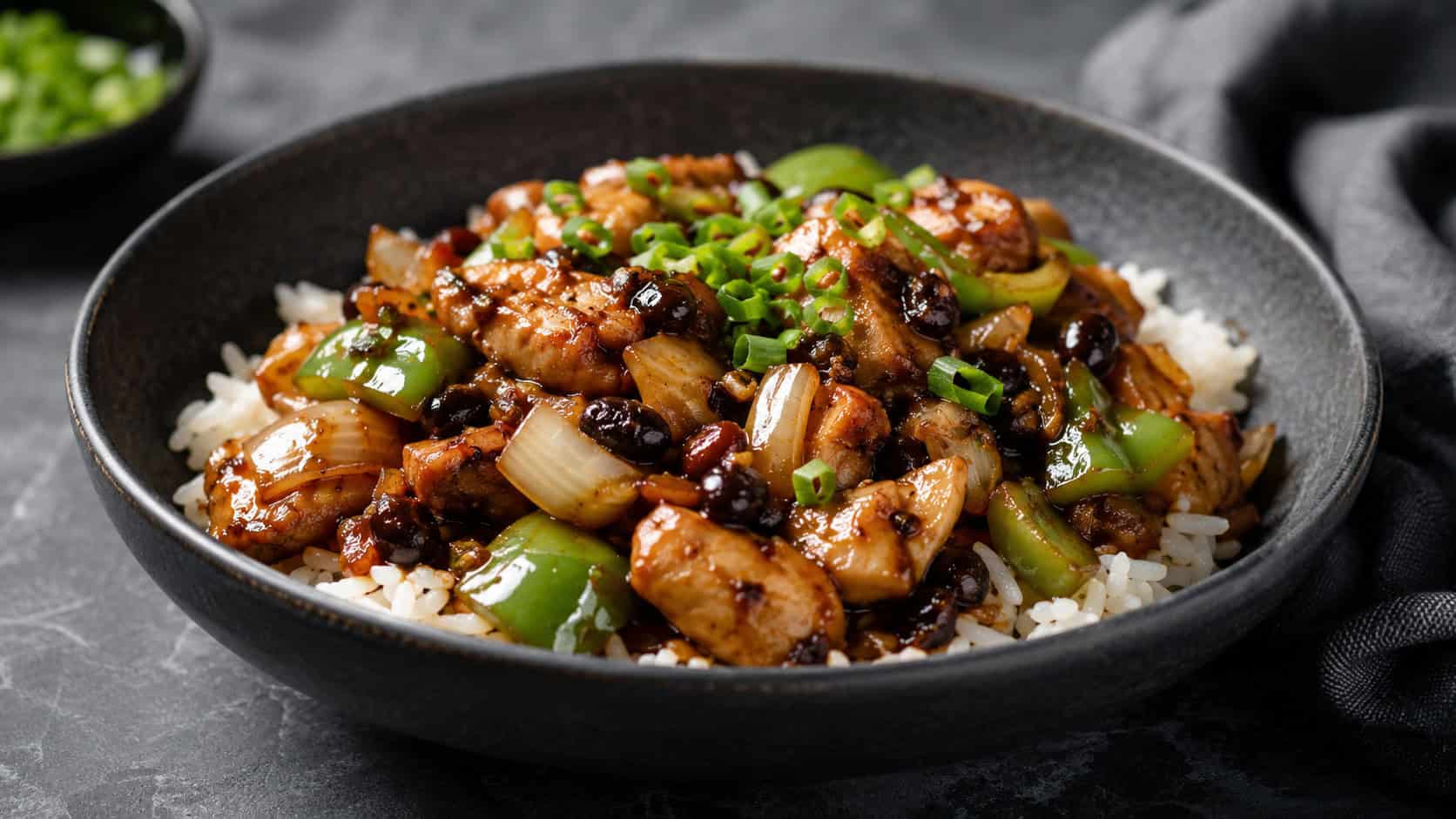 black bean chicken stir fry with green pepper and onion in a wide horizontal shot, dark savory sauce coating the chicken, served in a dark bowl over rice, topped with green onion, on a moody slate surface, natural light, shallow depth of field, landscape orientation