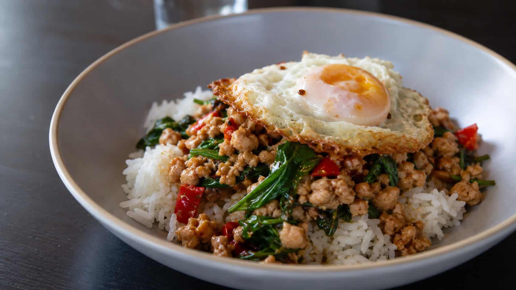 Thai basil chicken stir fry in a wide horizontal shot, savory minced chicken with wilted basil leaves and red chiles, served over jasmine rice in a shallow bowl, with a fried egg on top, on a dark table, natural light, shallow depth of field, landscape orientation