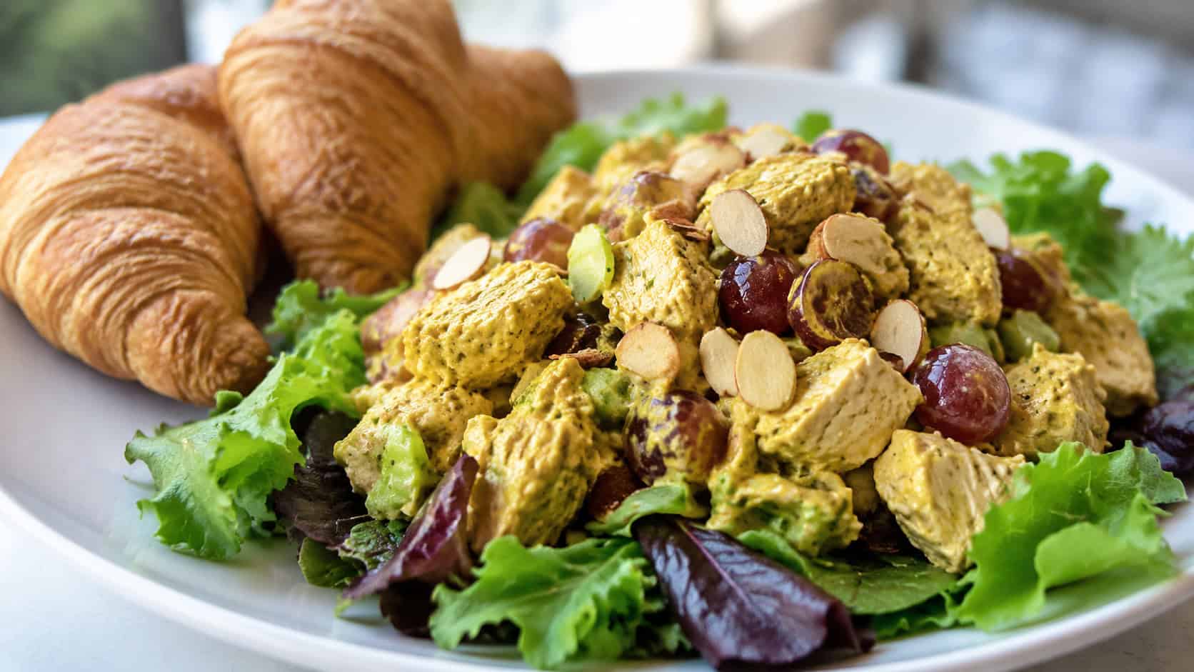 golden curried chicken salad with grapes and almonds on a bed of greens in a wide horizontal shot, croissants beside it, bright clean plate, soft natural light, shallow depth of field, landscape orientation