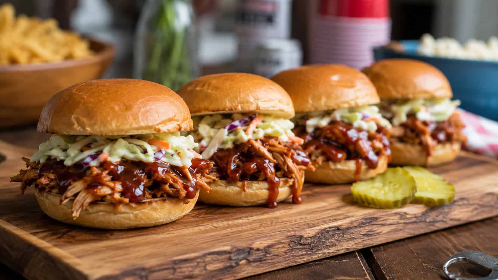 a row of barbecue chicken sliders topped with creamy coleslaw on a wooden board in a wide horizontal shot, glossy sauce visible, pickles on the side, casual party table, warm natural light, shallow depth of field, landscape orientation