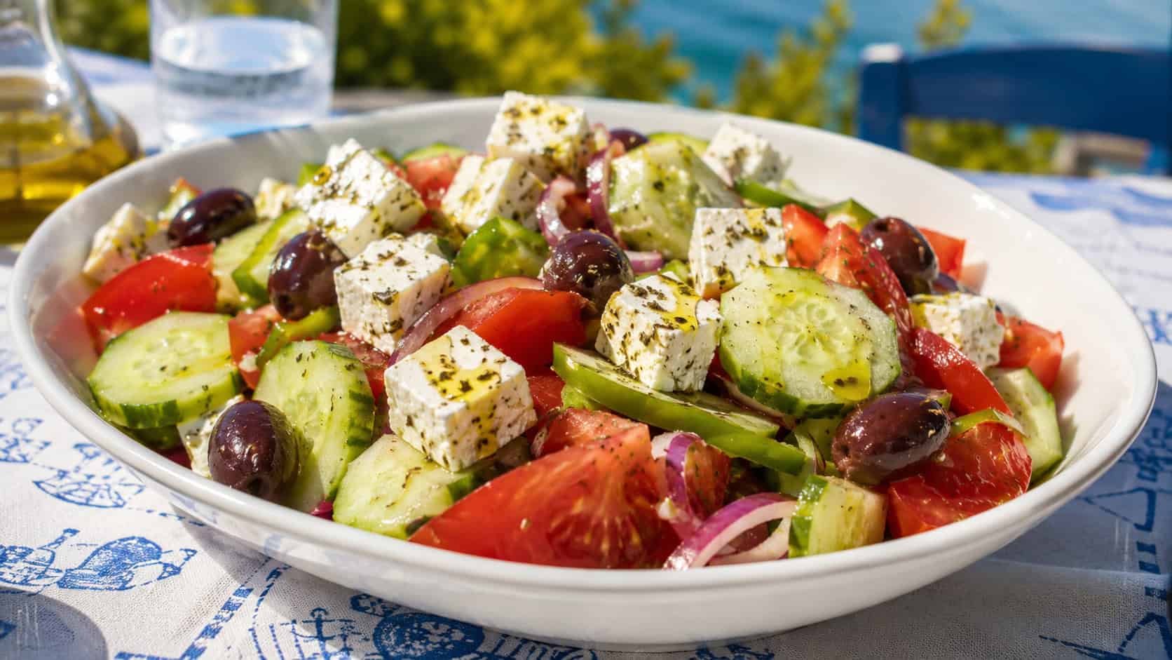 a colorful Greek salad with cucumbers, tomatoes, olives, and feta in a wide horizontal bowl, drizzled with olive oil, set on a sunny outdoor table, bright natural light, shallow depth of field, landscape orientation