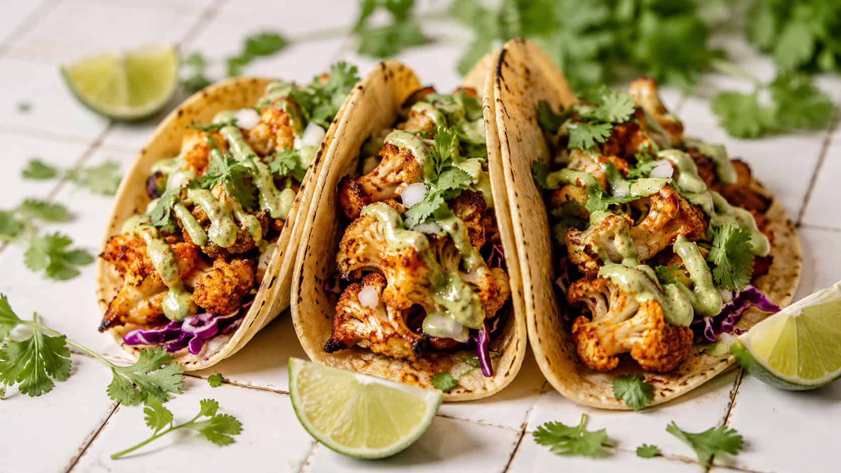 three cauliflower tacos with creamy avocado drizzle in a wide horizontal shot, lime wedges and cilantro scattered around, set on a bright tiled surface, natural light, shallow depth of field, landscape orientation