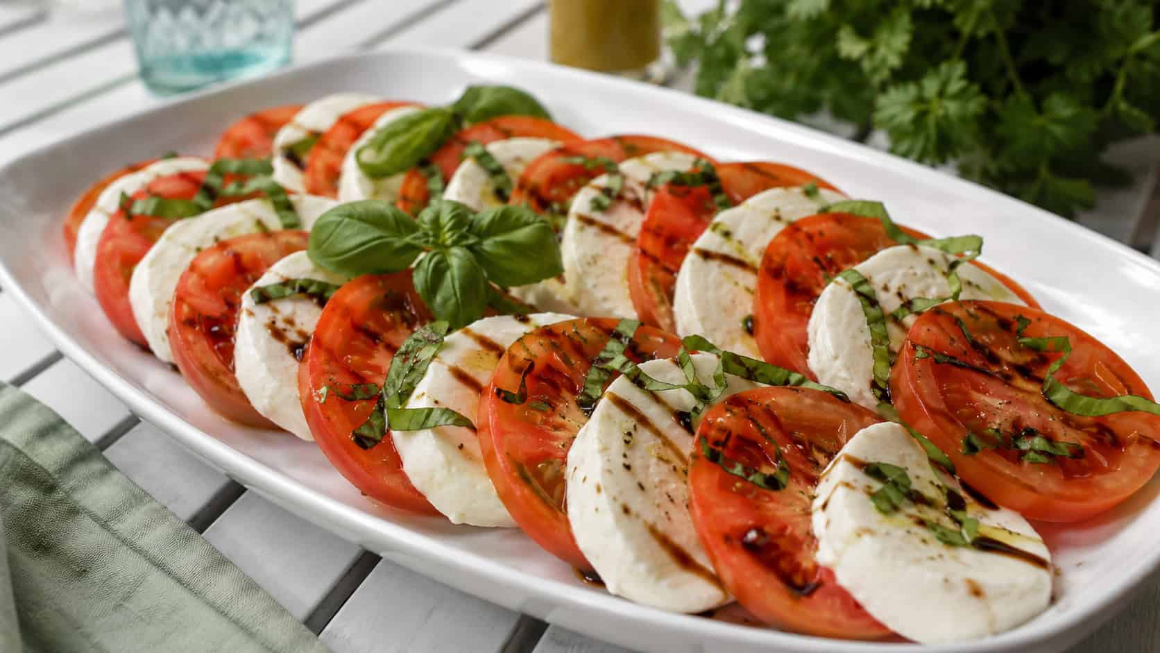 a caprese salad with alternating tomato and mozzarella slices in a wide horizontal platter, fresh basil and balsamic drizzle on top, set on a bright outdoor table, natural light, shallow depth of field, landscape orientation