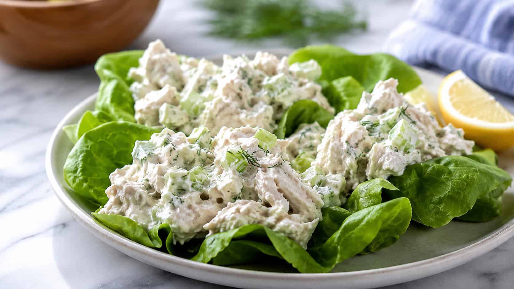 creamy Greek yogurt chicken salad with flecks of celery and dill served on lettuce leaves on a plate, lemon wedge nearby, on a bright table, wide horizontal shot, natural light, shallow depth of field, landscape orientation