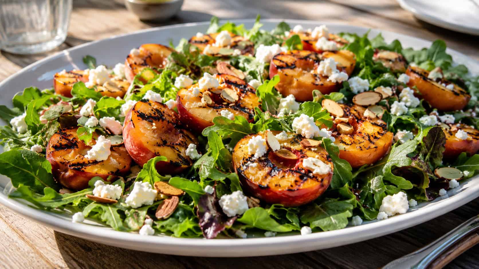 a grilled peach salad with charred peach halves, crumbled white feta, and toasted almonds over fresh greens on a large platter, on a sunlit table, wide horizontal shot, natural light, shallow depth of field, landscape orientation