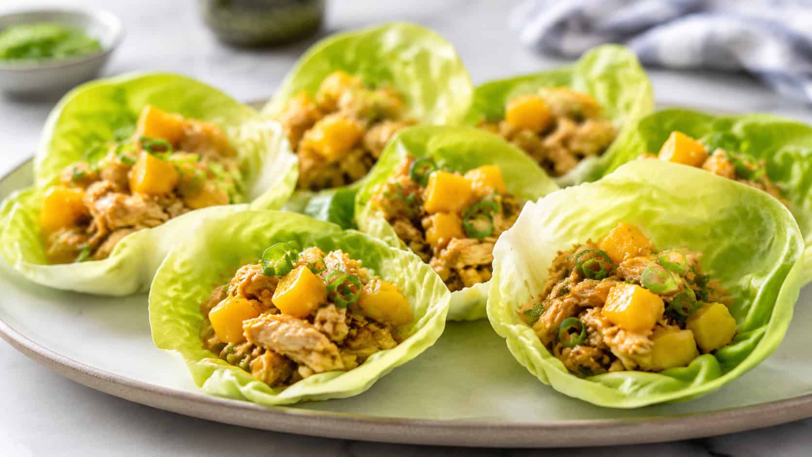 butter lettuce cups filled with savory chicken and bright orange mango, garnished with green onion, arranged on a platter, on a bright table, wide horizontal shot, natural light, shallow depth of field, landscape orientation