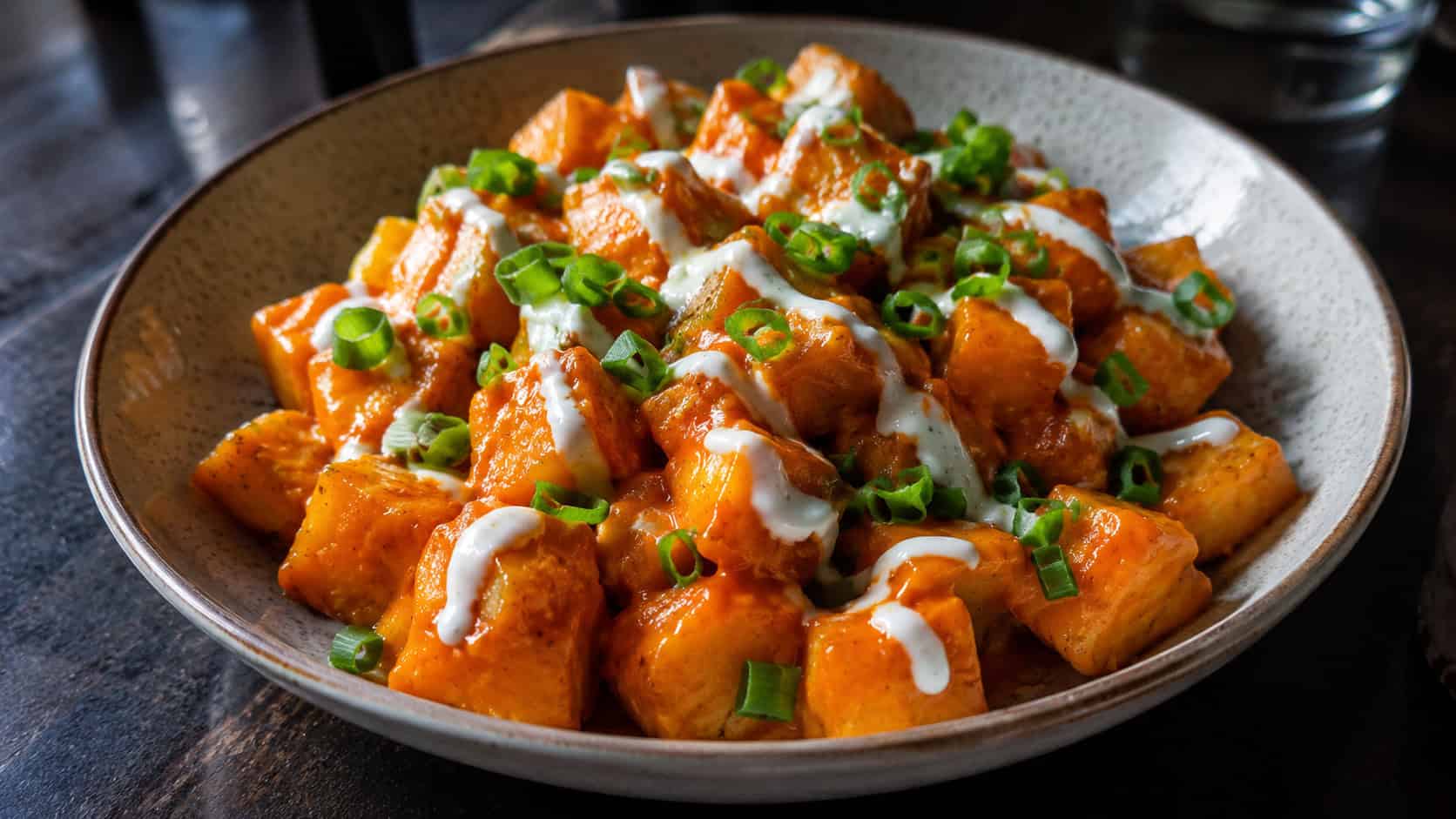 buffalo ranch potato bites in a wide horizontal shot, glossy orange-coated cubes drizzled with white ranch and green onion, in a shallow bowl on a dark table, warm natural light, shallow depth of field, landscape orientation